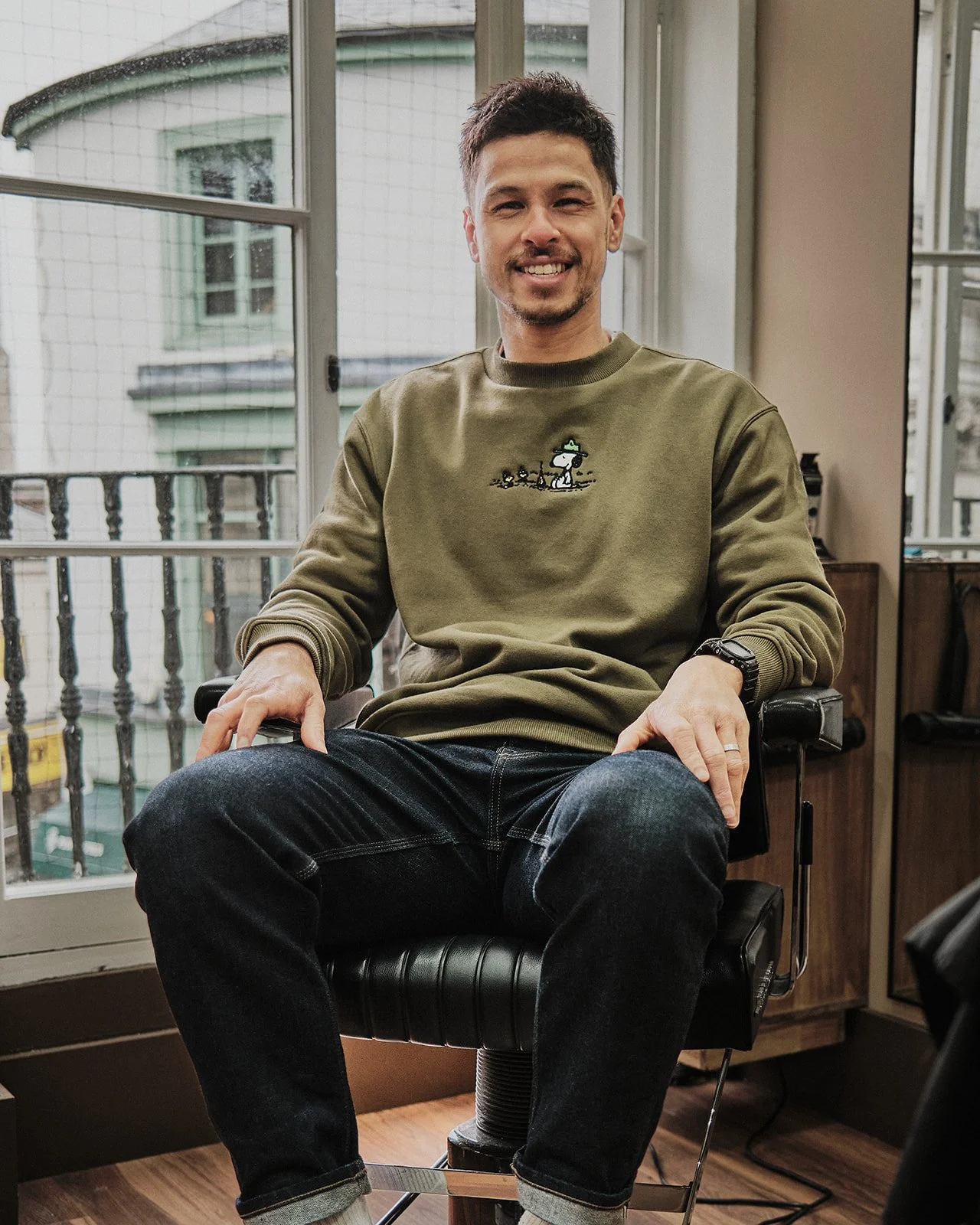 Man sitting in a chair by a large window, smiling, wearing a green sweatshirt with a Snoopy design and dark jeans.