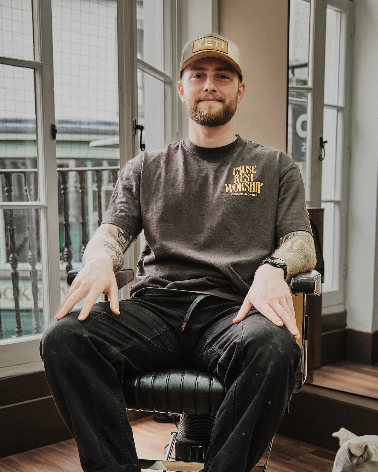 A man with a beard and tattoos sitting in a barber shop chair, wearing a baseball cap and a T-shirt with the words 'Pause Rest Worship' on it, and looking at the camera.