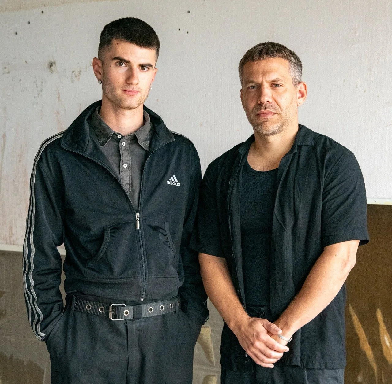 Two men standing side by side against a plain wall, both with serious expressions, wearing black shirts and casual black jackets.