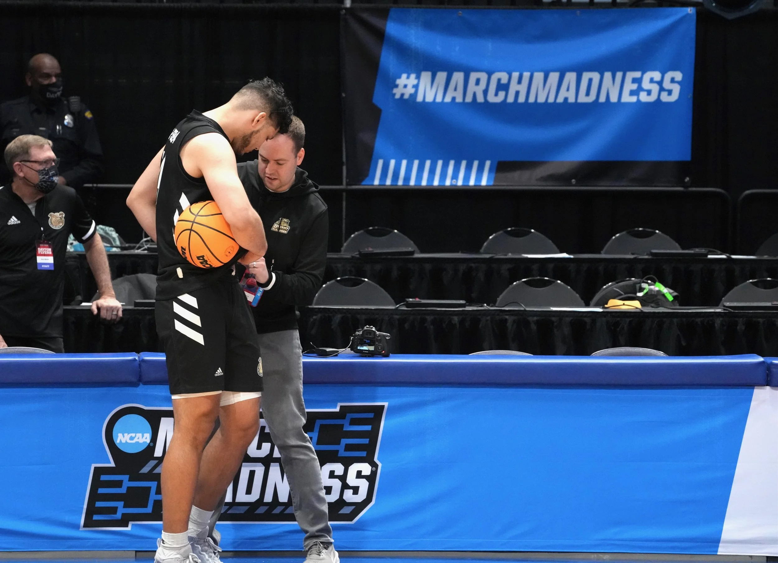 A basketball player and videographer standing on a court with a blue NCAA March Madness banner in the background.