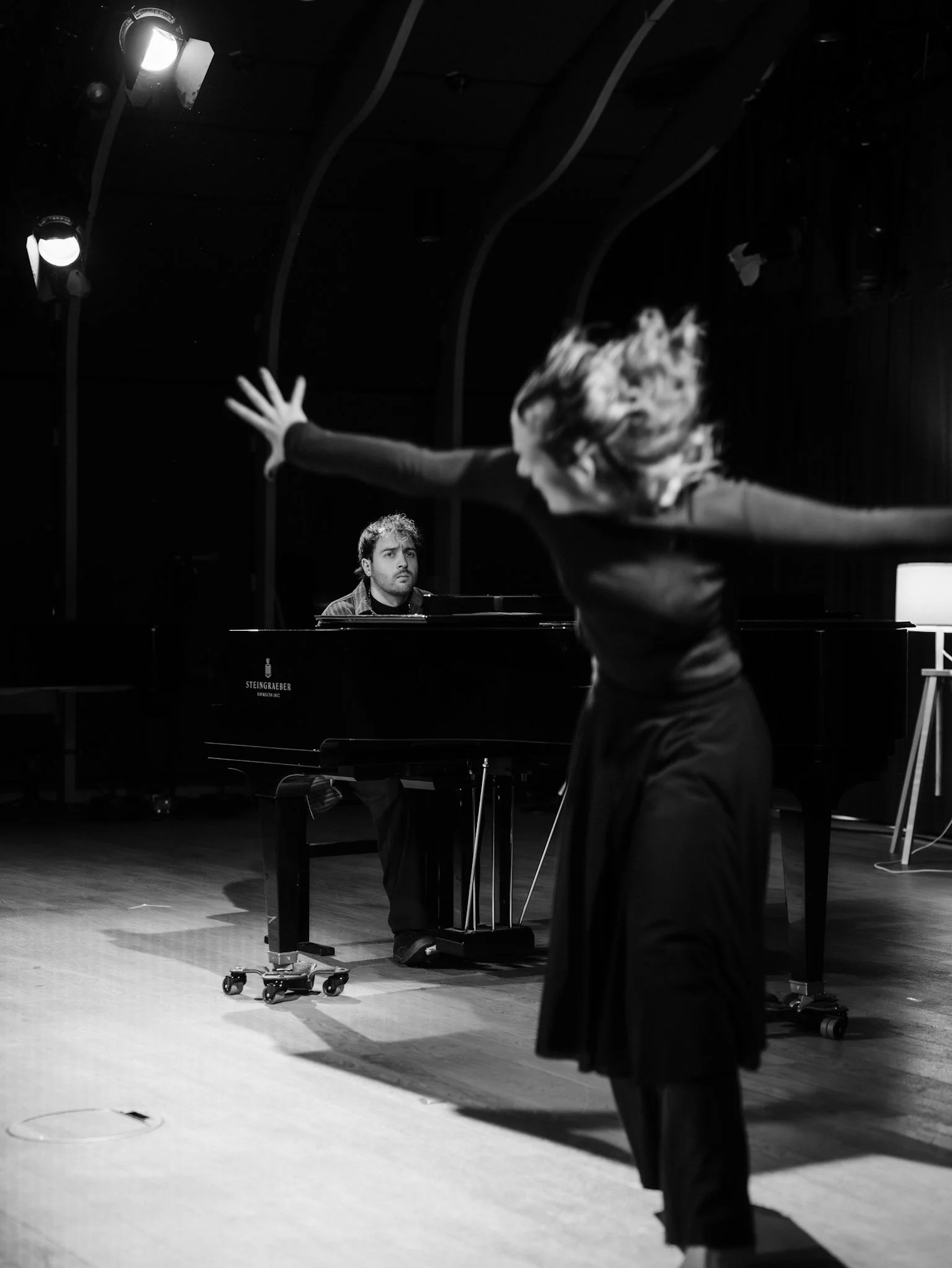 A woman with her arms extended and hair flying is performing a dance or expressive movement in front of a man sitting at a grand piano, in a dimly lit performance space.