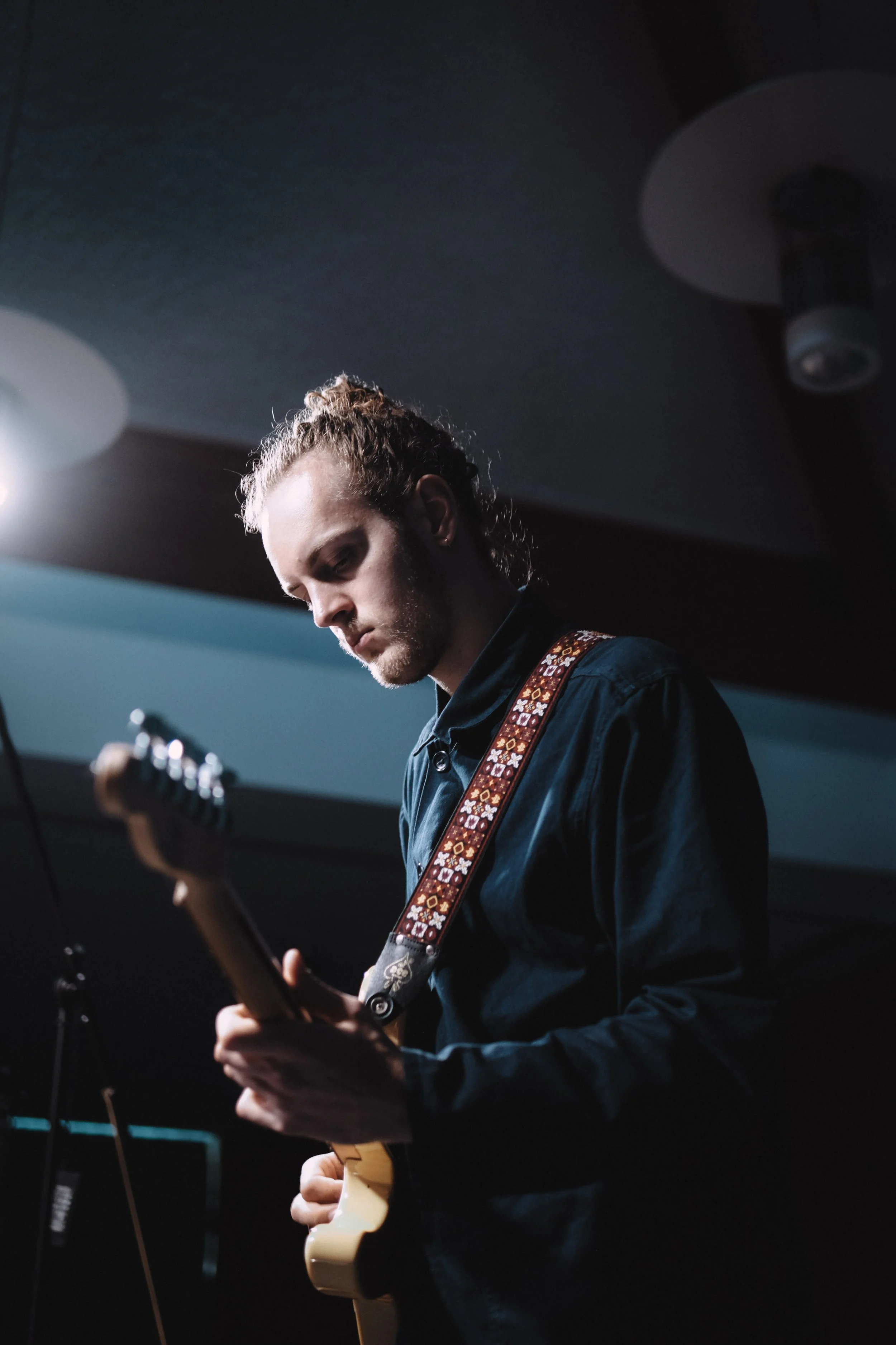 A young man with curly hair playing an electric guitar on stage, focused on his performance, with stage lights overhead.