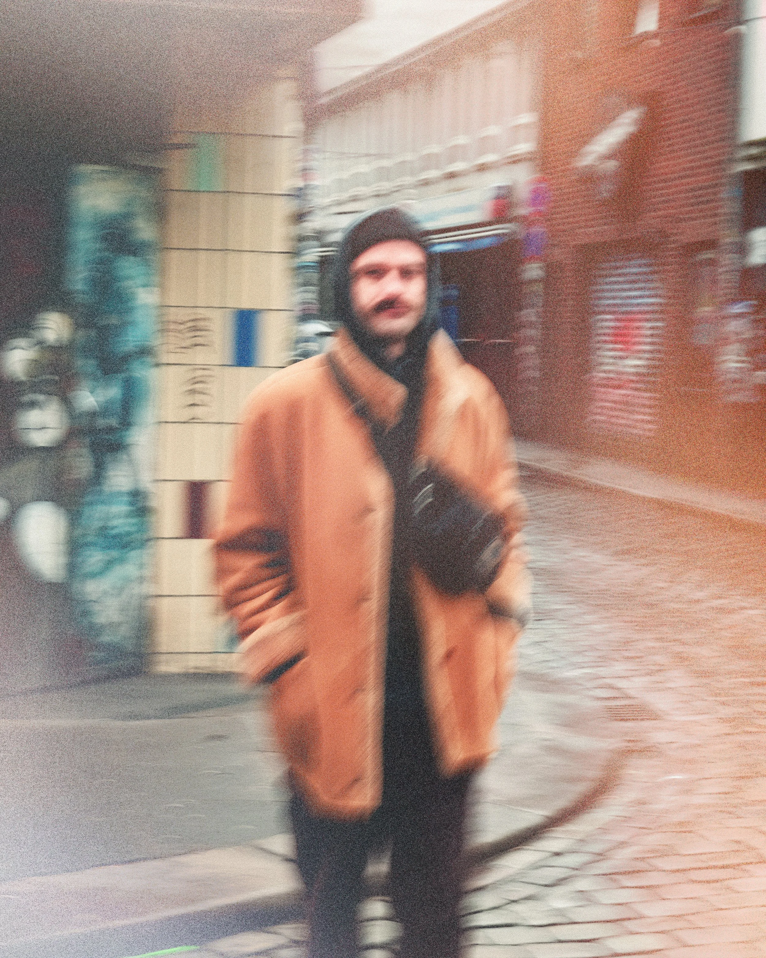 Blurred image of a man wearing a brown jacket and black beanie, walking on a city street at night with blurred storefronts and signs in the background.