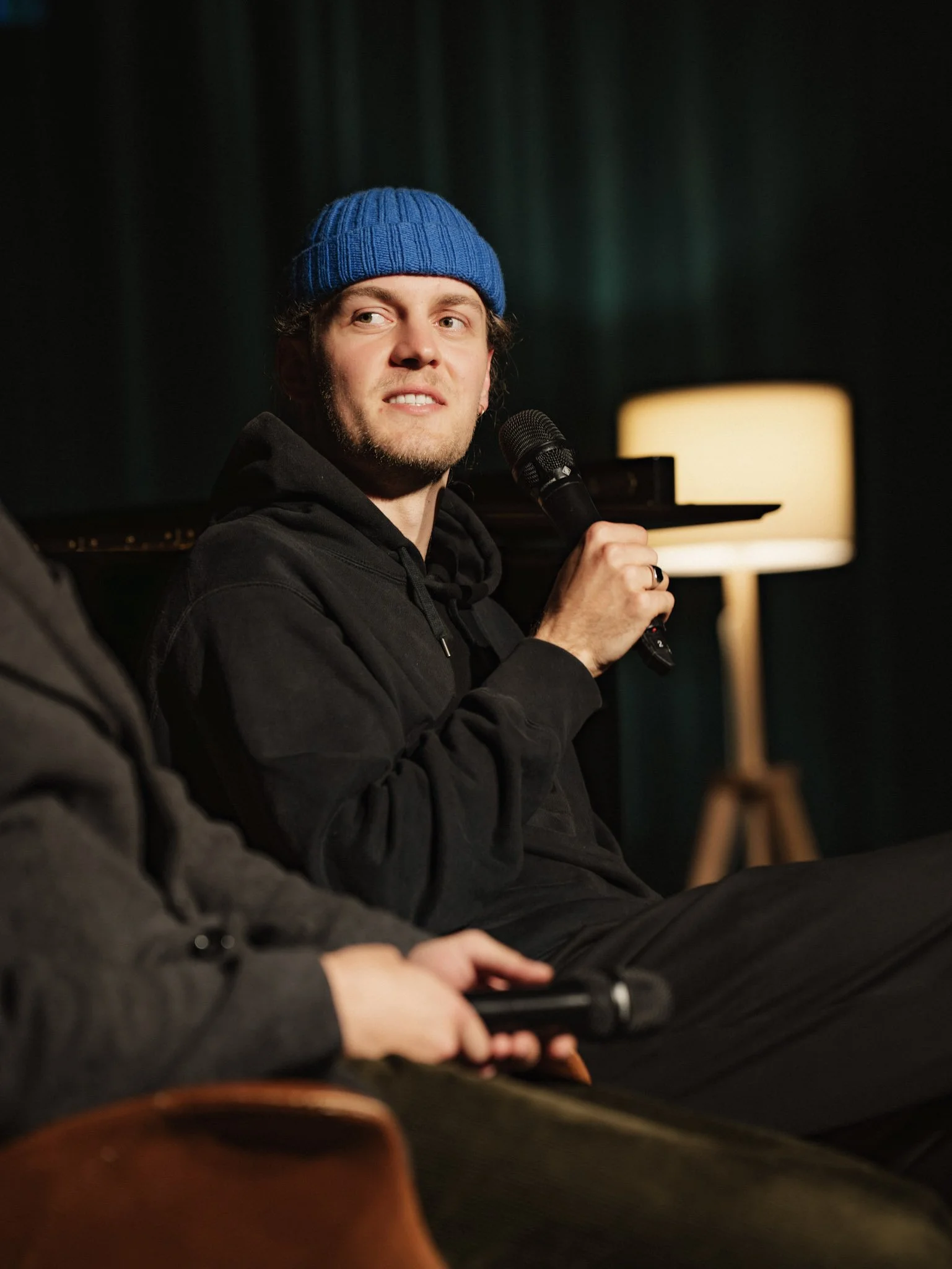 A man wearing a blue knit cap holding a microphone. He is seated indoors with a black background and a beige floor lamp in the background.