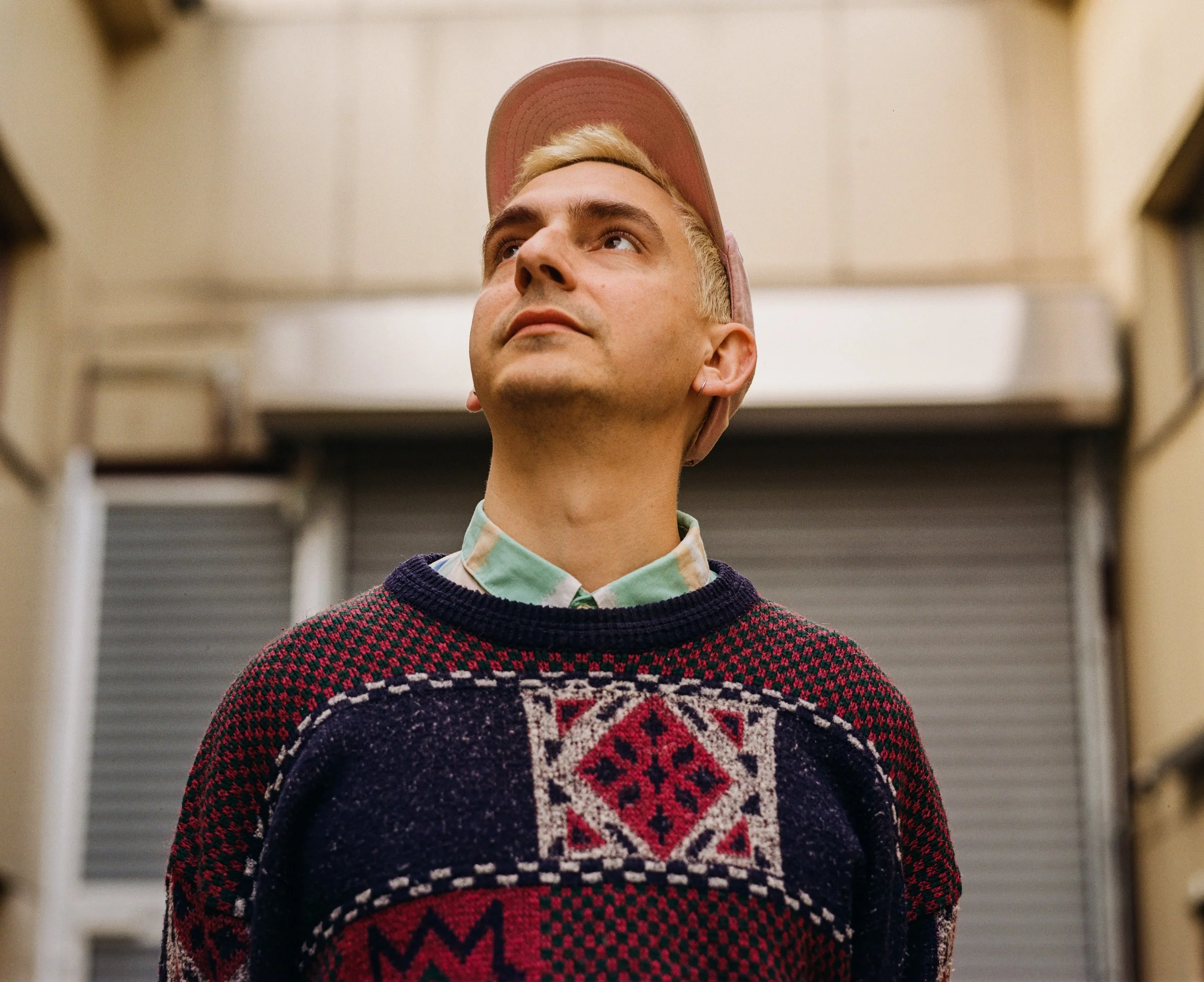 A young man wearing a pink cap, a pastel green shirt, and a festive Christmas sweater, standing in a hallway, looking up.