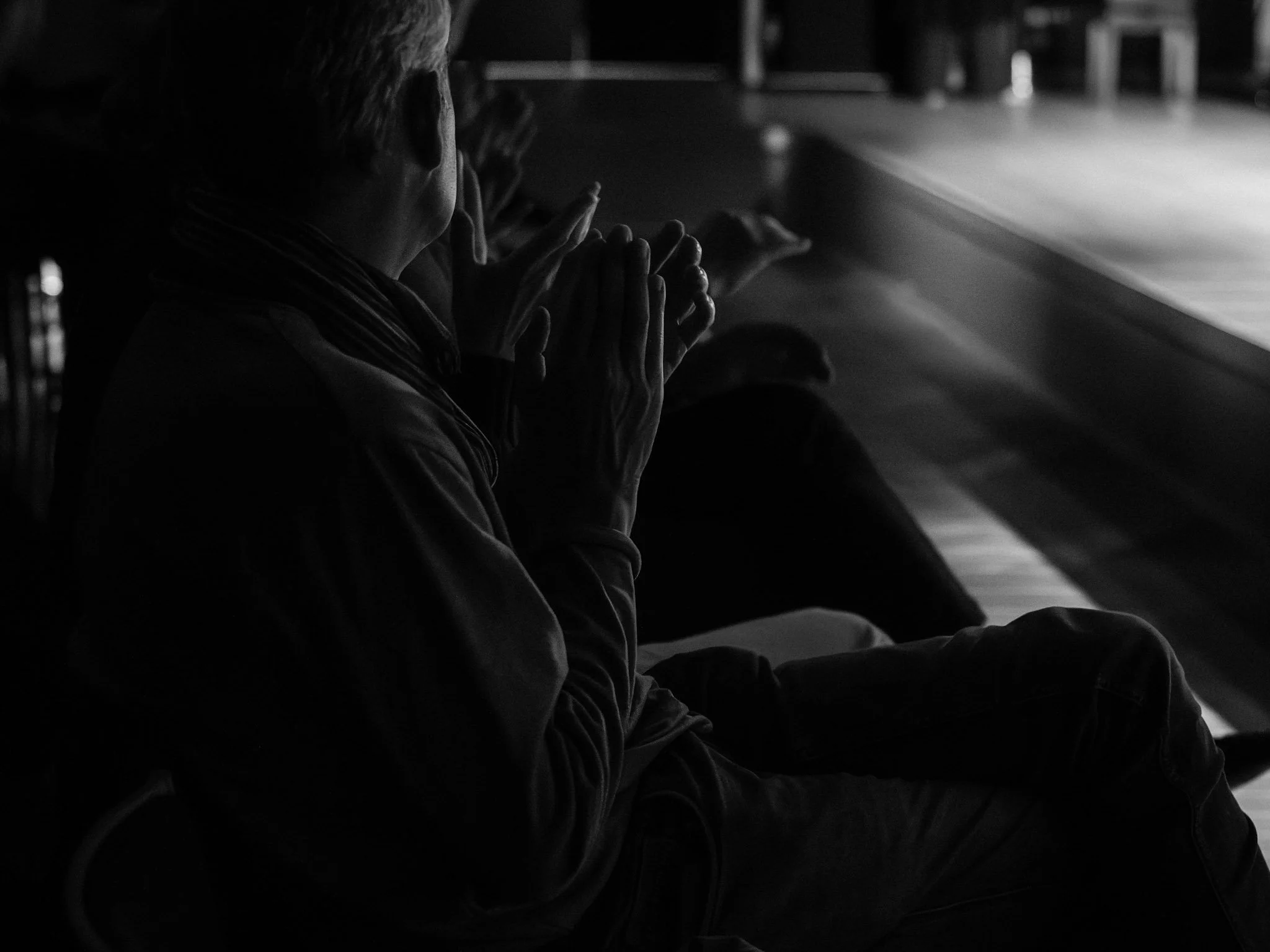 A person with short hair sitting in a darkened room, with hands clasped as if praying, facing a lighted area or screen.
