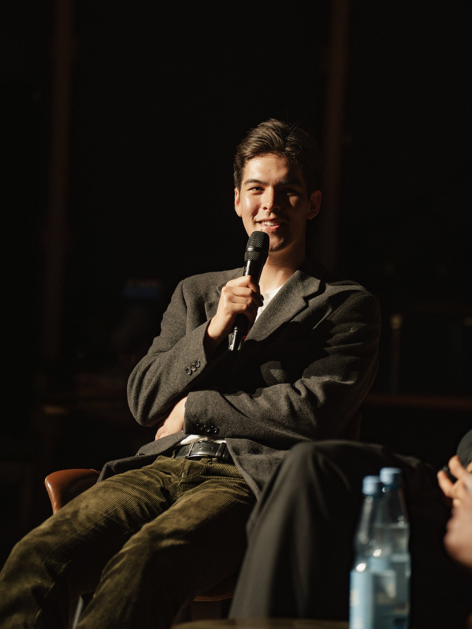 A young man with short dark hair and light skin holding a microphone, dressed in a dark blazer and green pants, sitting on a brown chair in a dimly lit room with a dark background.