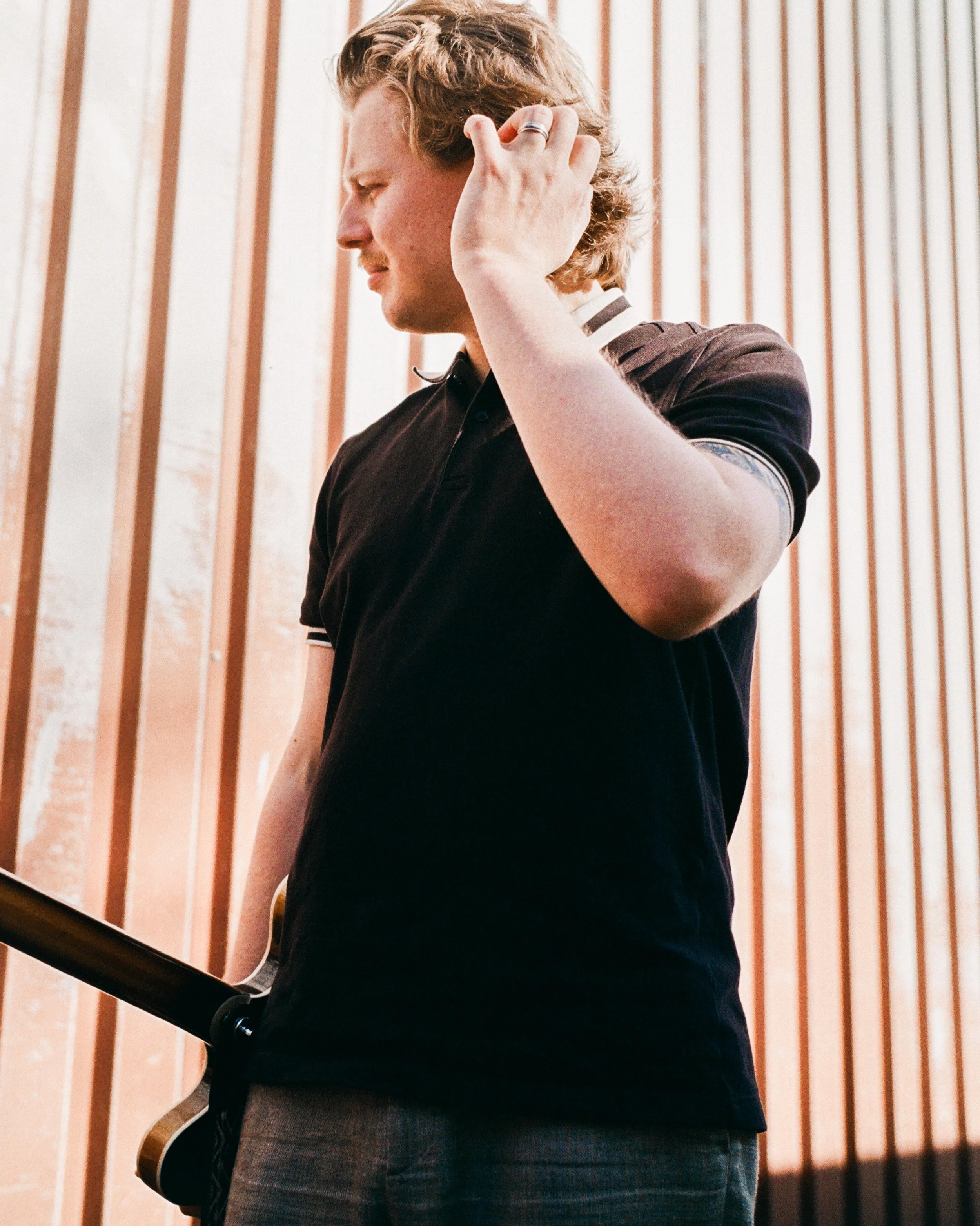 A person with curly blonde hair wearing a black shirt with a striped collar holding an acoustic guitar with a wooden body and white pickguard, standing against a background of vertical wooden panels.