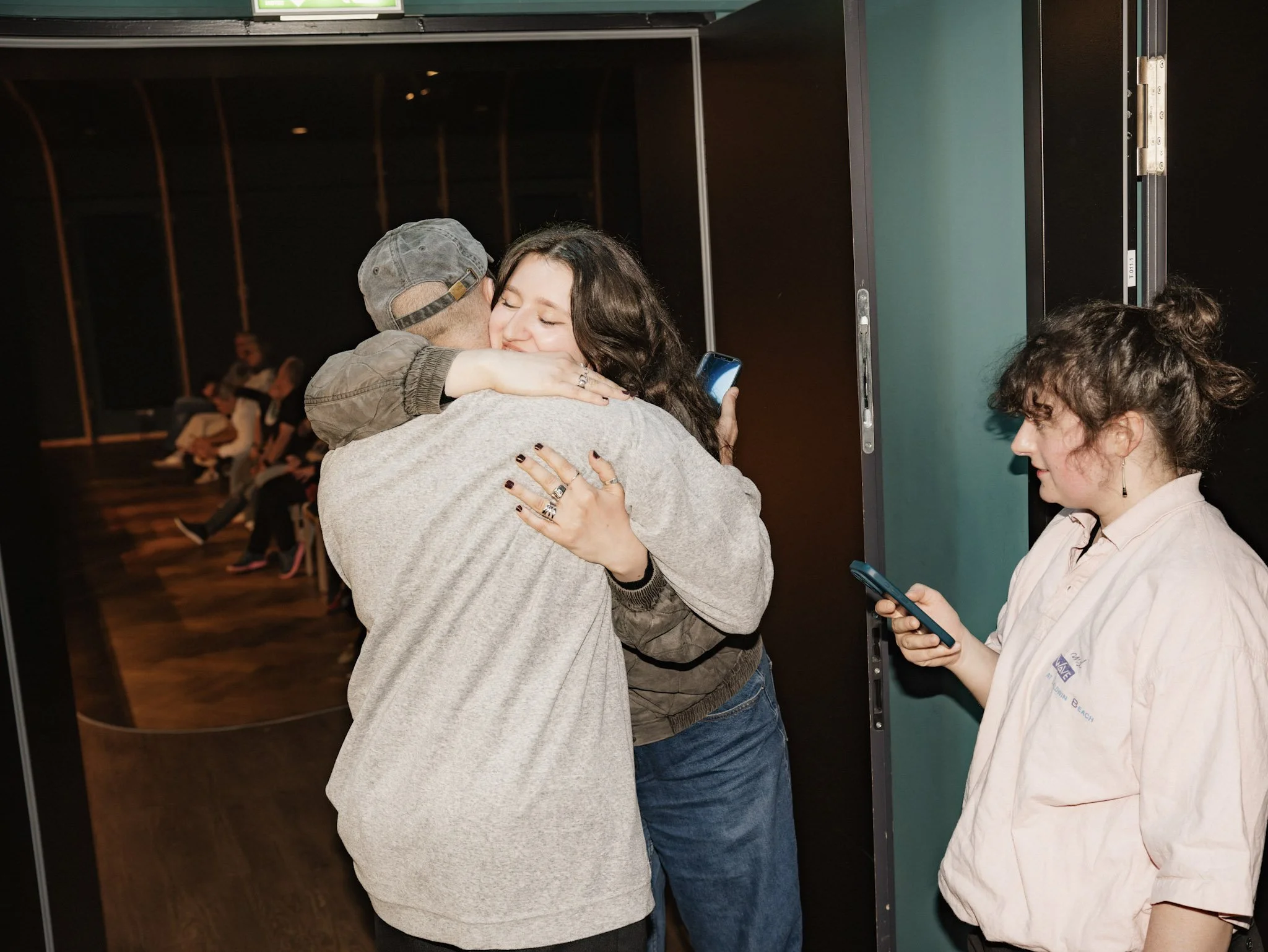 Two people hugging while one person holds a smartphone, standing near a green door, with other people sitting in the background.