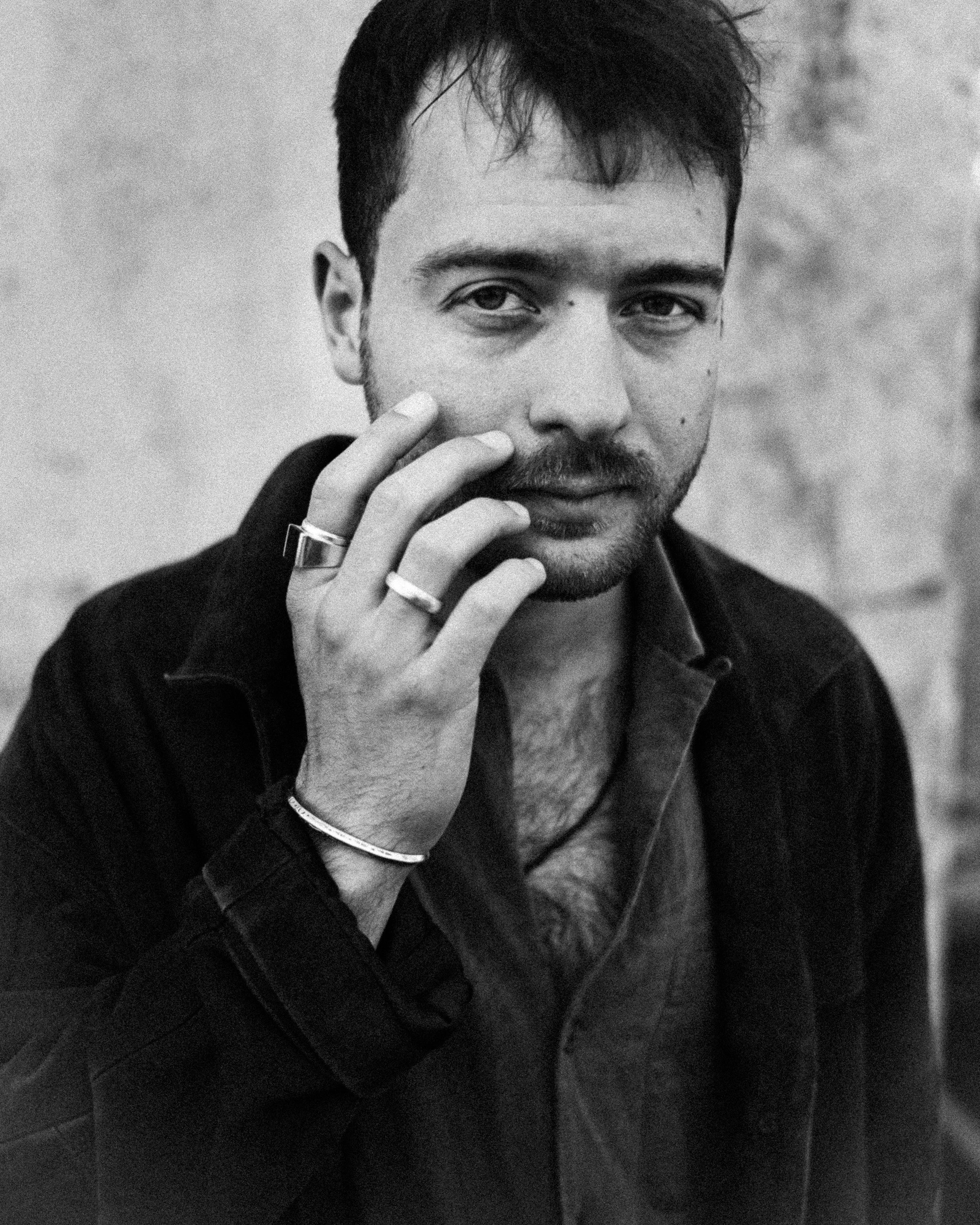 A black-and-white portrait of a man with short dark hair and a beard, holding his fingers near his face, wearing rings and a bracelet, with a blurred background.