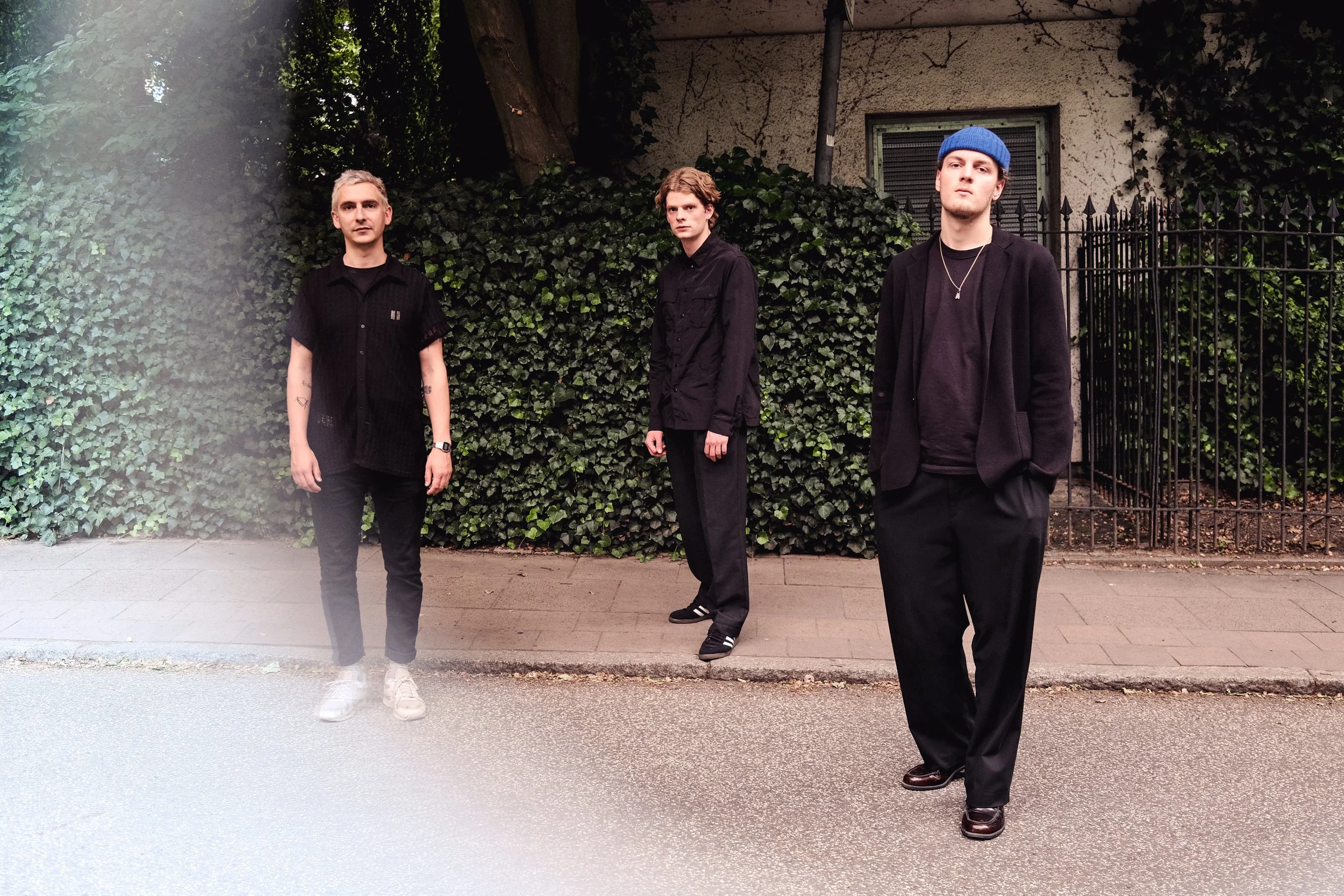 Three young men standing on a sidewalk in front of a wall covered with green ivy, with a gated fence visible on the right. They are dressed in black with casual and stylish outfits, looking directly at the camera.