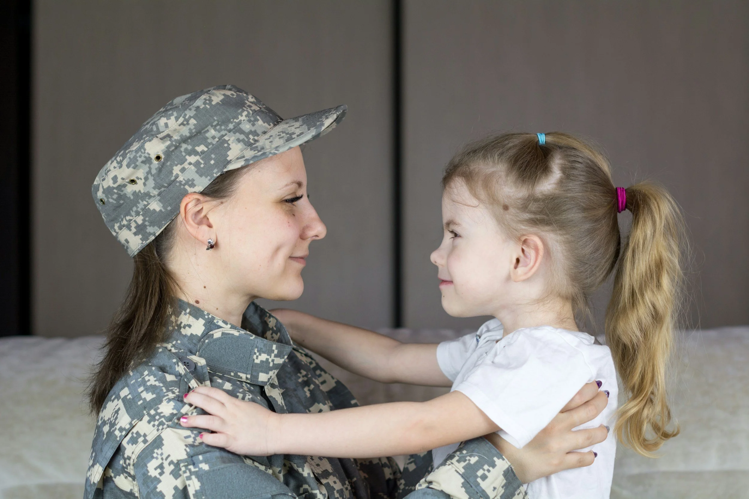 A woman in military uniform with a camouflage pattern faces a young girl. The woman is smiling and holding the girl, who is smiling back. The girl has long blonde hair tied in pigtails with colorful hair ties. They are looking at each other, and their foreheads are touching.