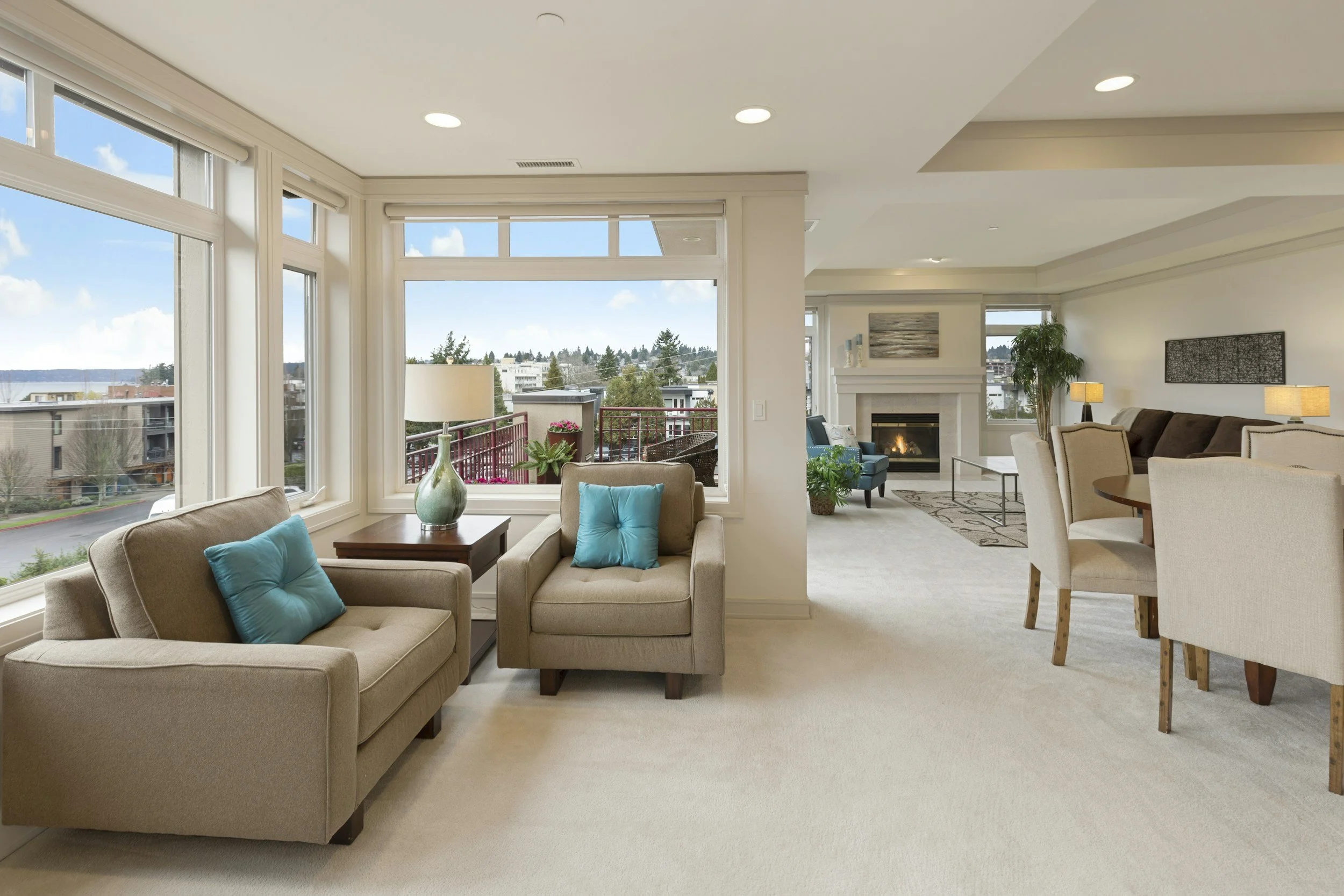 Living room with large windows, beige sofas with blue pillows, a fireplace, a dining area, potted plants, and a balcony view of a neighborhood and trees.