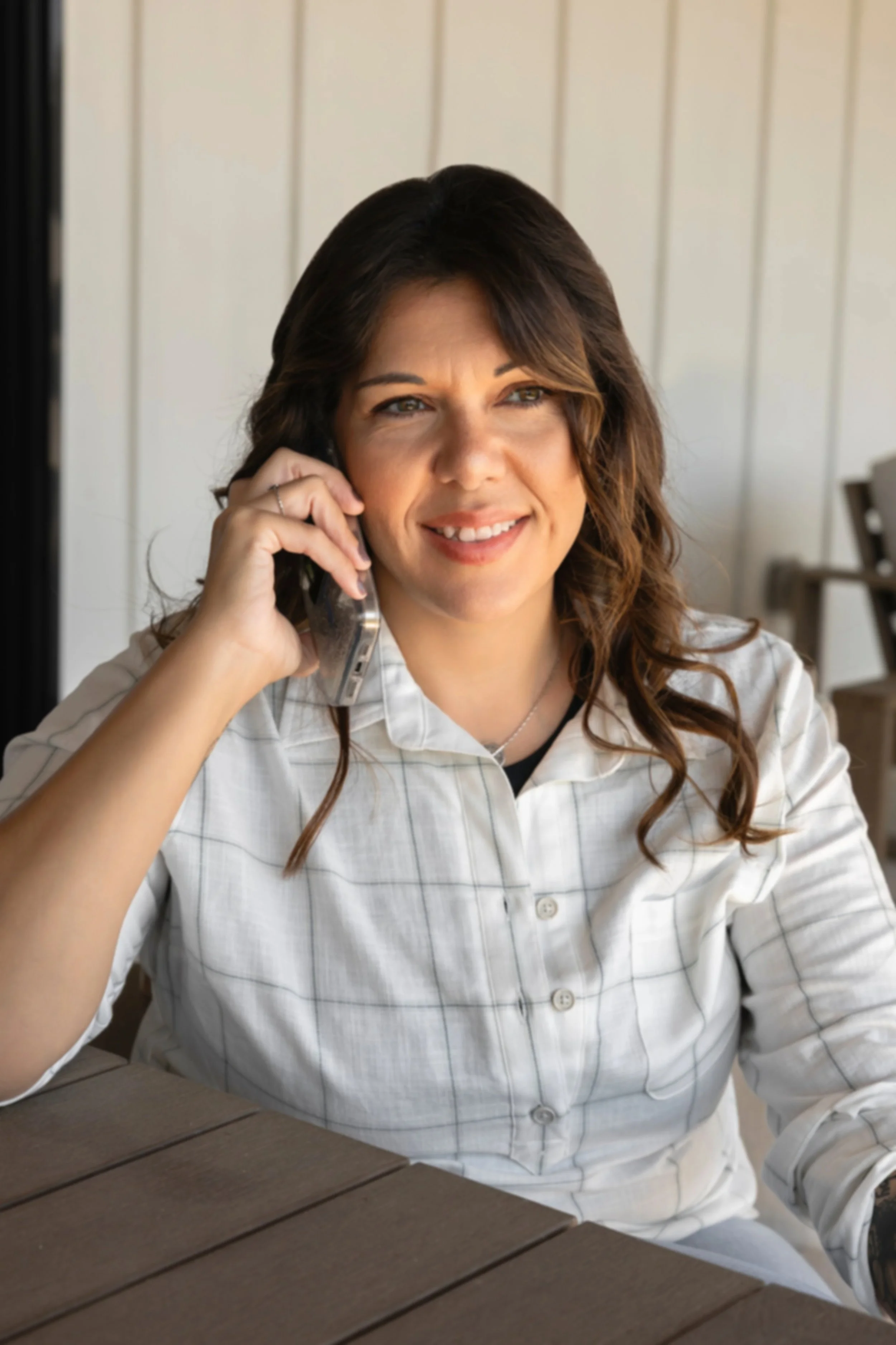 Woman with wavy brown hair talking on a landline phone, wearing a white checked shirt, sitting at a wooden table with a white paneled wall in the background.