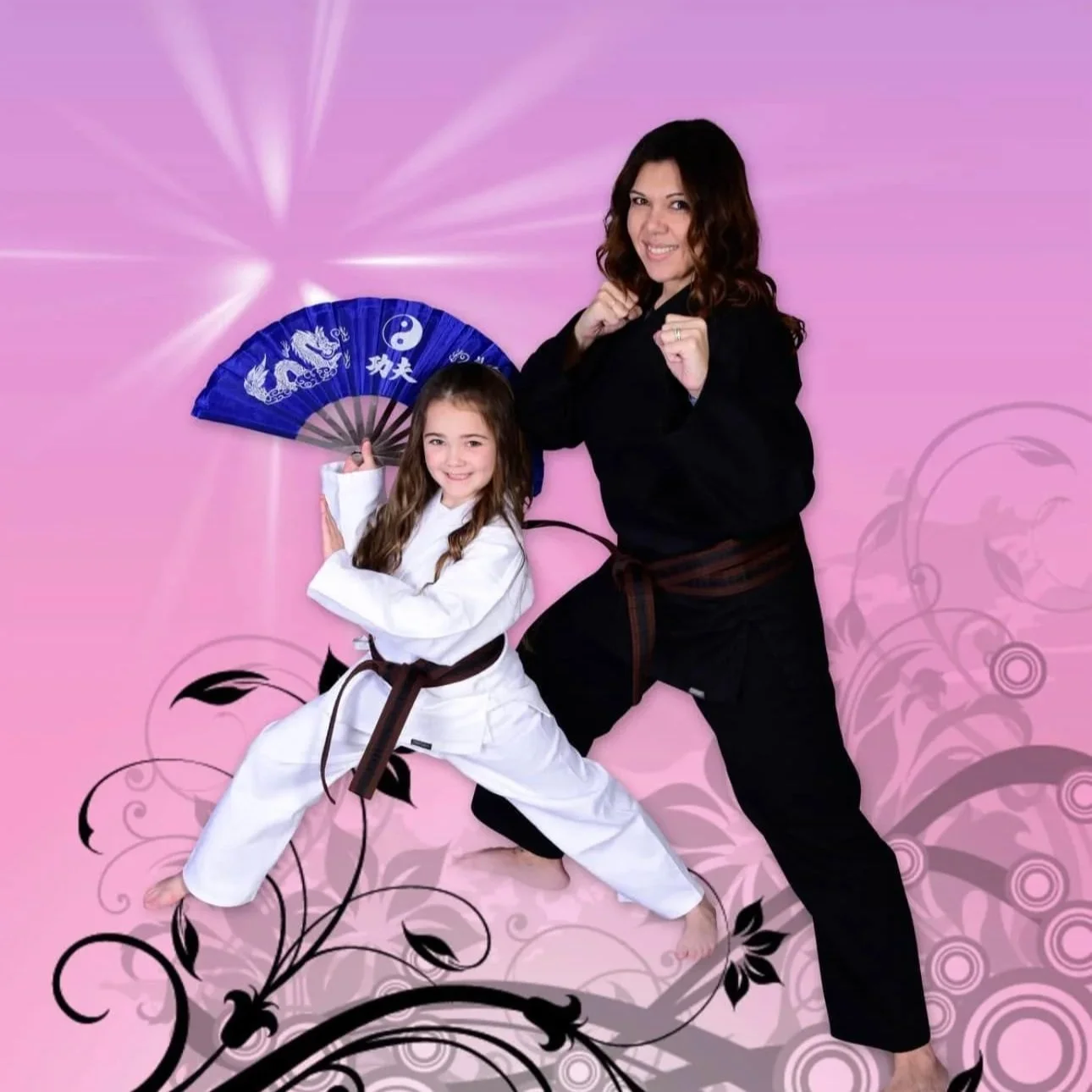 A woman and a young girl dressed in martial arts uniforms pose in front of a pink background with black floral designs. The girl holds a blue martial arts fan, smiling, while the woman strikes a fighting pose.
