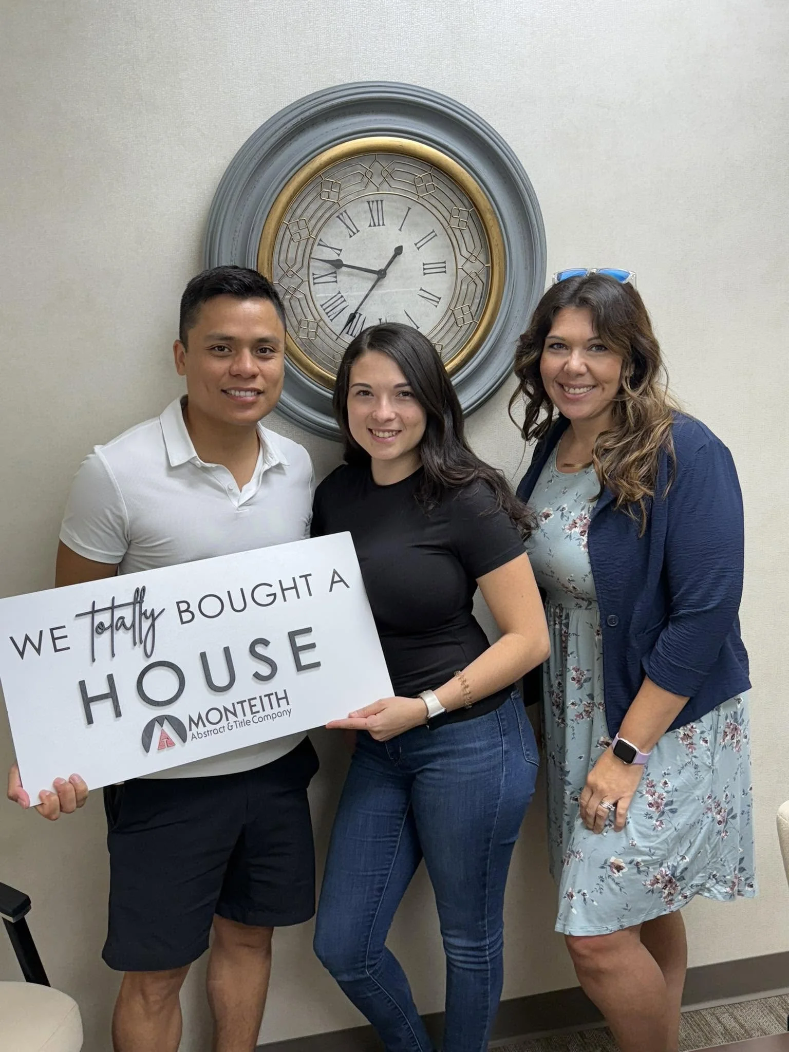 Three women standing together in front of a large wall clock, holding a sign that says 'WE TALLY BOUGHT A HOUSE'. They are smiling and dressed casually, with one woman wearing glasses on her head and another wearing a smartwatch.