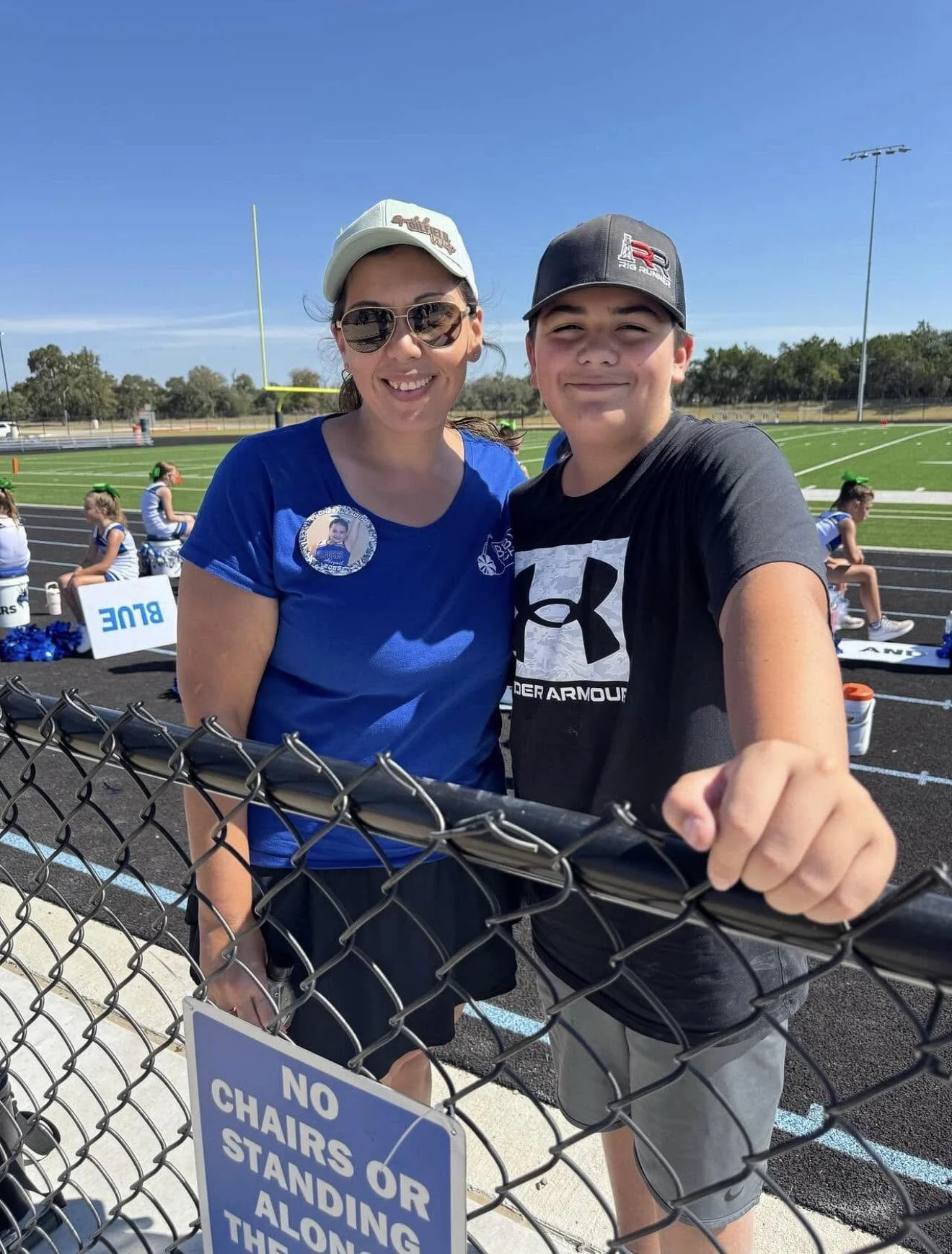 A woman and a boy smiling and taking a selfie at an outdoor football field. The woman is wearing sunglasses, a light-colored cap, and a blue shirt with a badge. The boy is wearing a black Under Armour shirt and a black cap. There are cheerleaders sitting in the background and a sign on the fence that says 'No Chairs or Standing Along the'.