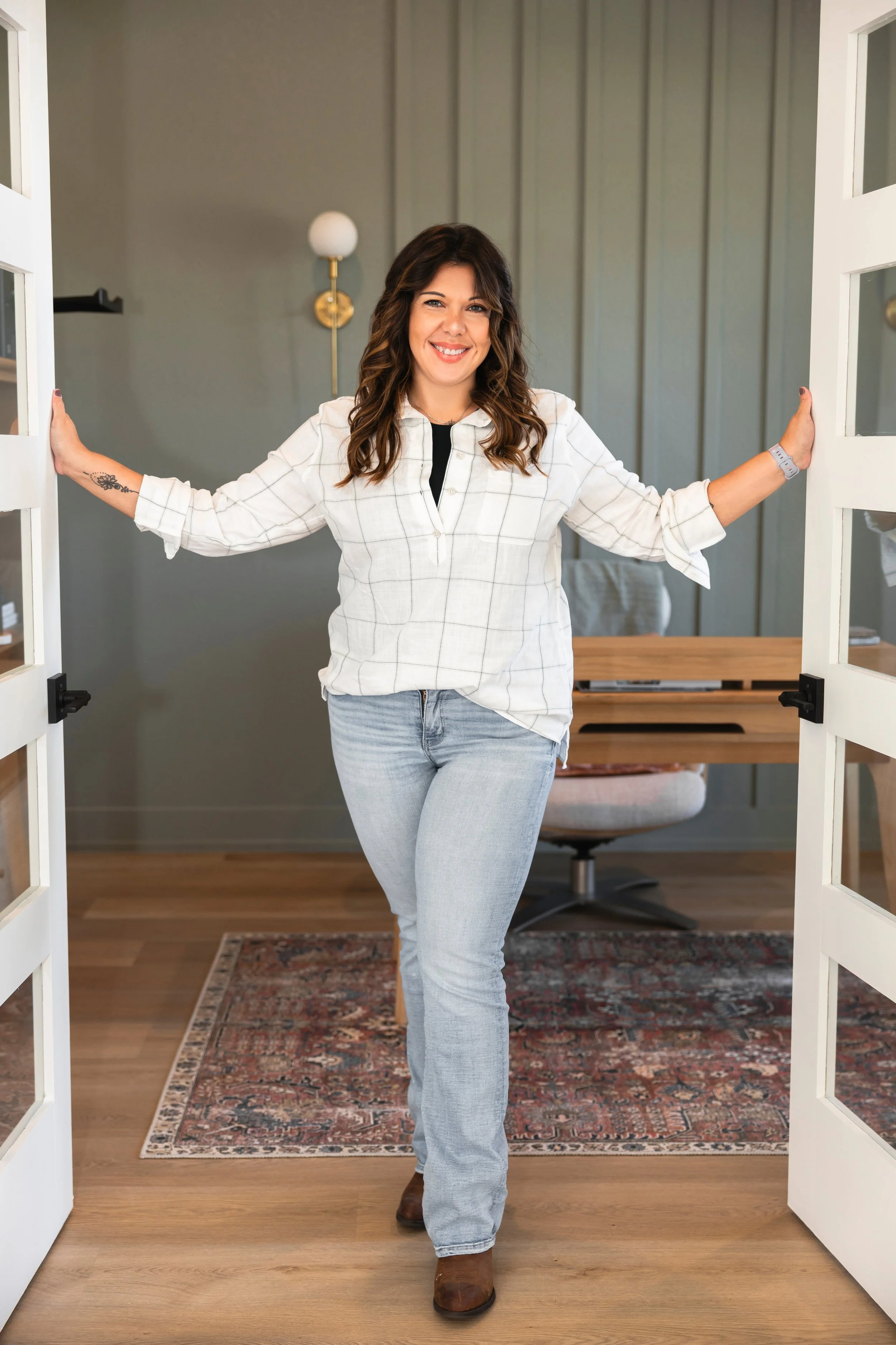A woman with dark brown wavy hair, wearing a white plaid shirt, light blue jeans, and brown boots, standing in a doorway with a smile. The background features a gray-green accent wall, a wall lamp, a wooden table, and a Persian-style rug on the wooden floor.