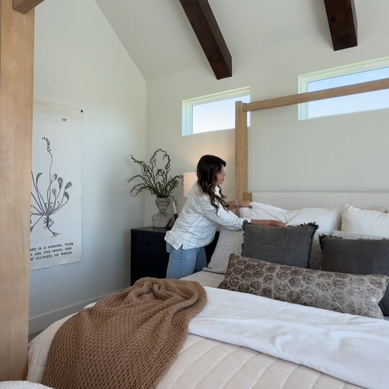 A woman changing pillows on a bed in a bedroom with vaulted ceiling, white walls, middle window, and decorative pillows.