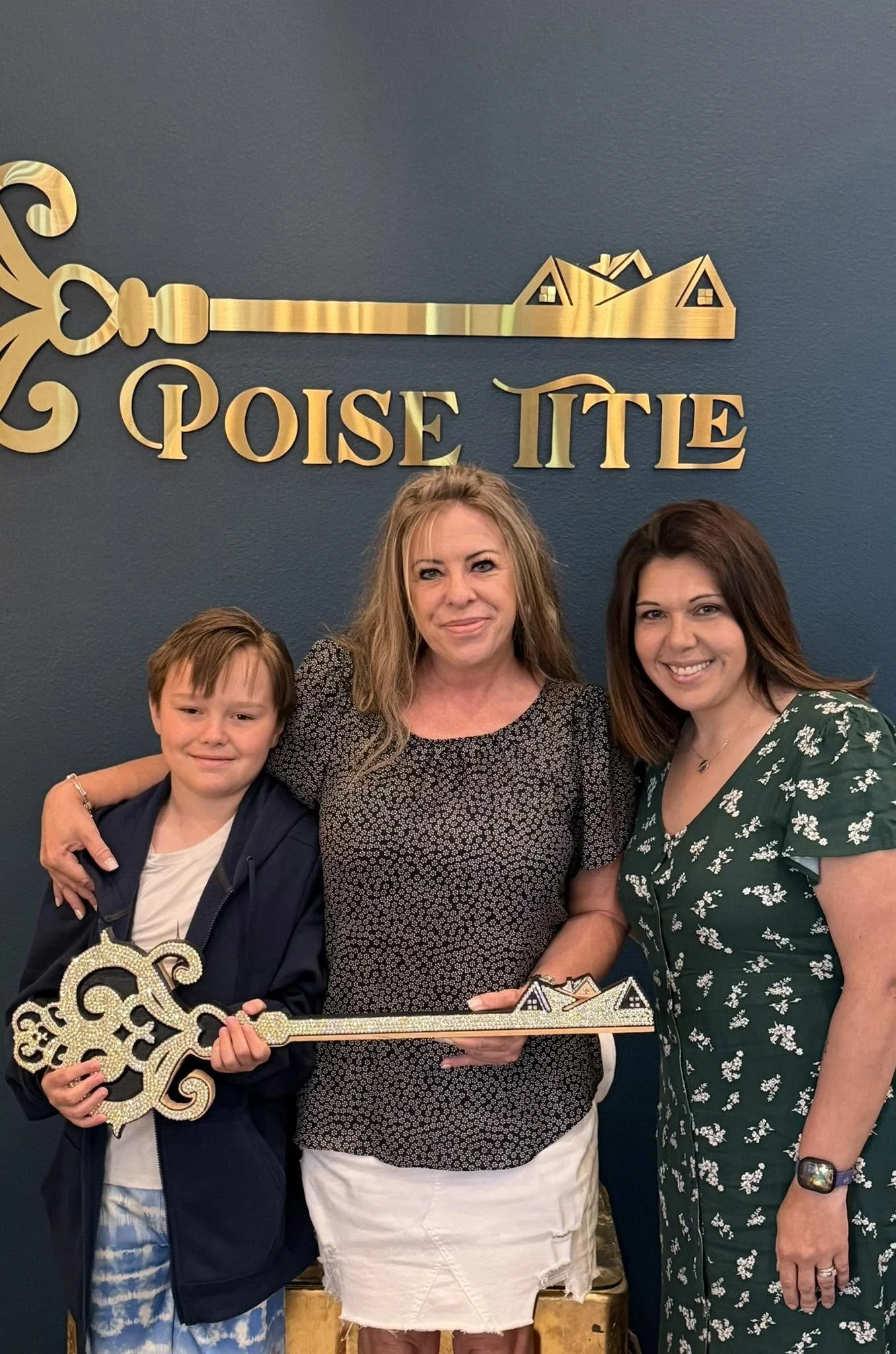 Three women and a young boy pose for a photo in front of a dark blue wall with gold signage that reads 'Poise TUE' above a stylized house. The woman in the middle is holding a large, ornate, gold-colored key and a set of house-shaped decorations.