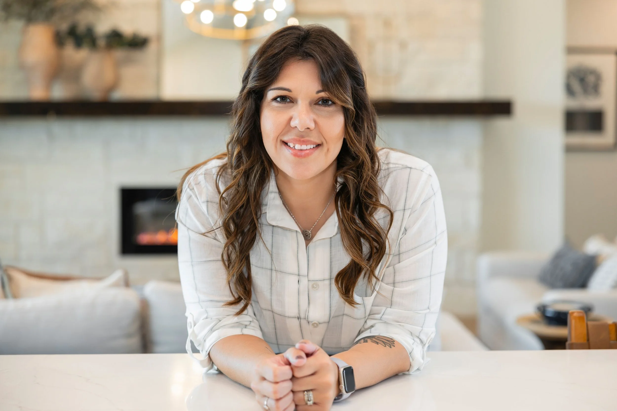 A woman with wavy brown hair smiling at the camera, sitting at a white table in a cozy, modern kitchen with a fireplace in the background.