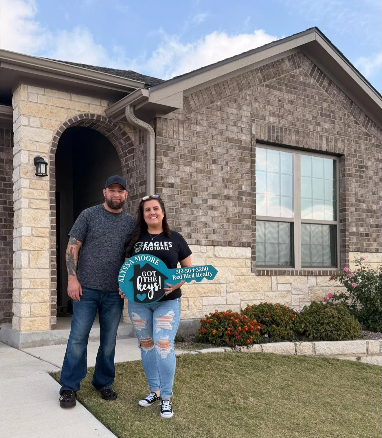 A couple standing in front of a house with a brick exterior, holding a sign that says 'Alyssa Moore Got the Keys! 512-564-3260 Red Bird Realty'. The man has a beard, black cap, and tattoos on his arm, and the woman has long dark hair, wearing a black Eagles football shirt and ripped jeans.
