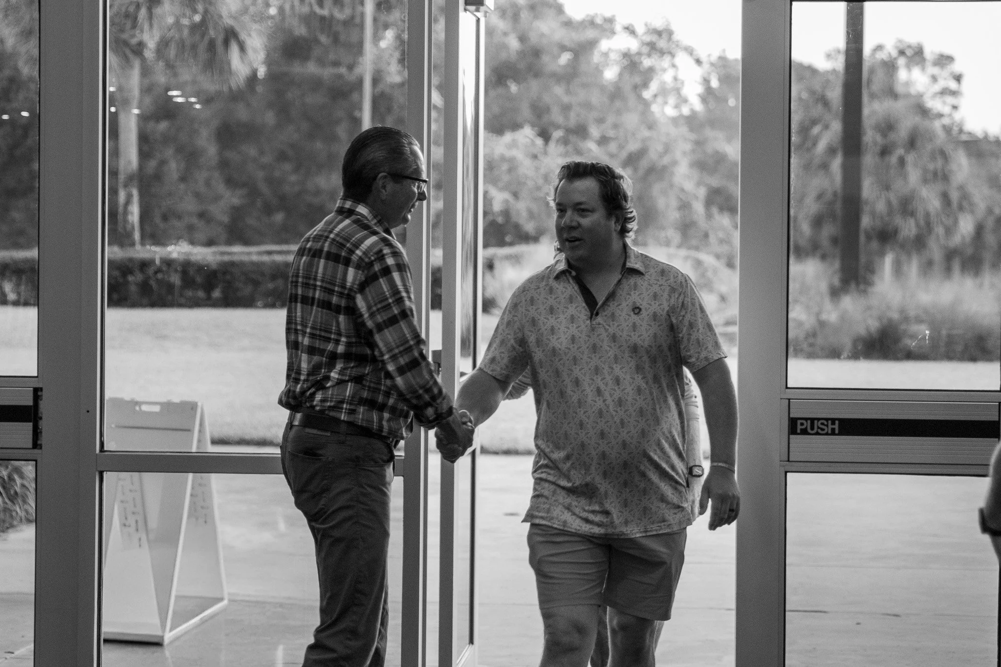 Two men shake hands as they exit a building through glass doors, with a park and trees visible outside.
