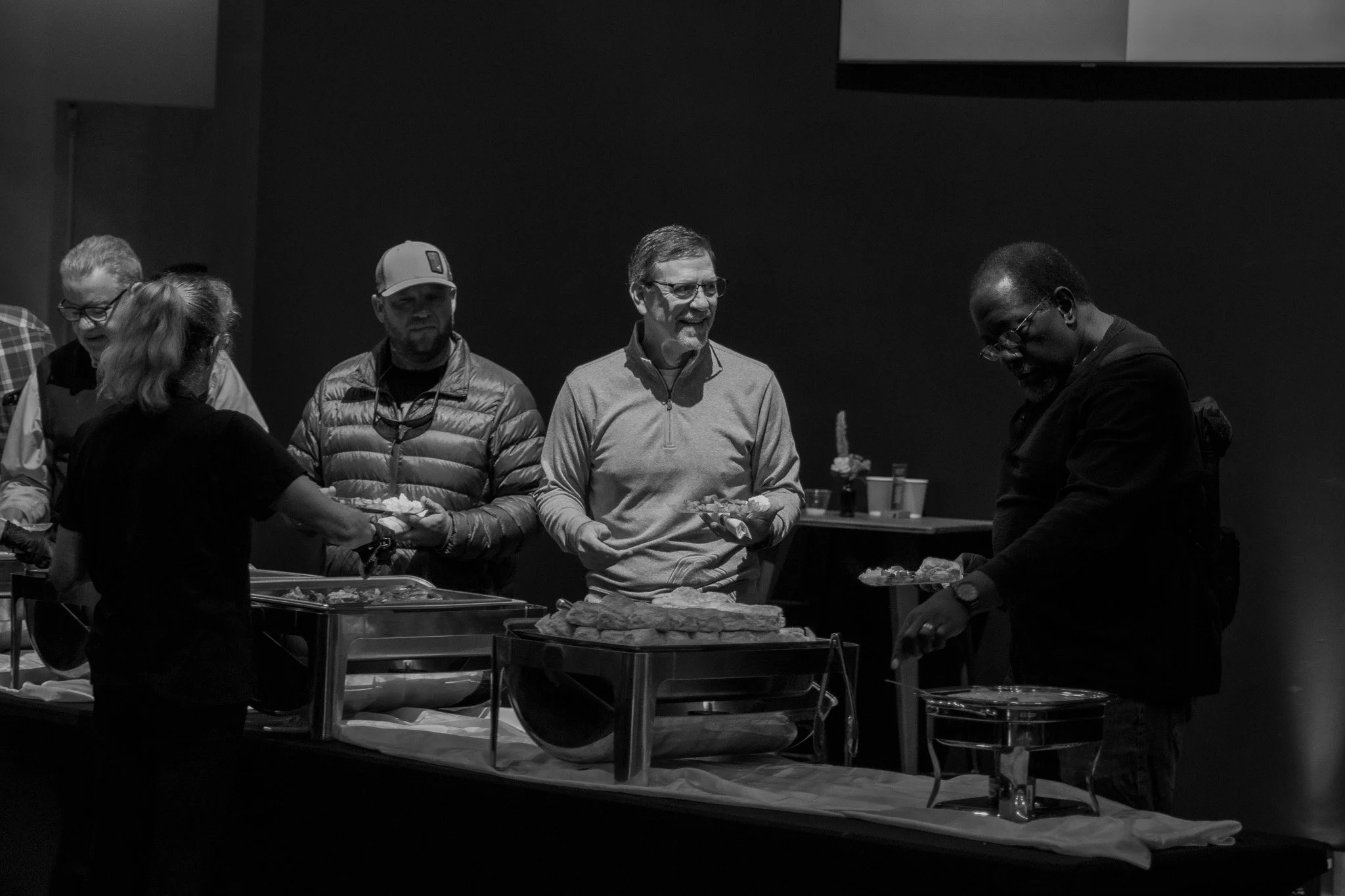 Several men in line at a buffet with plates of food, engaging in conversation.