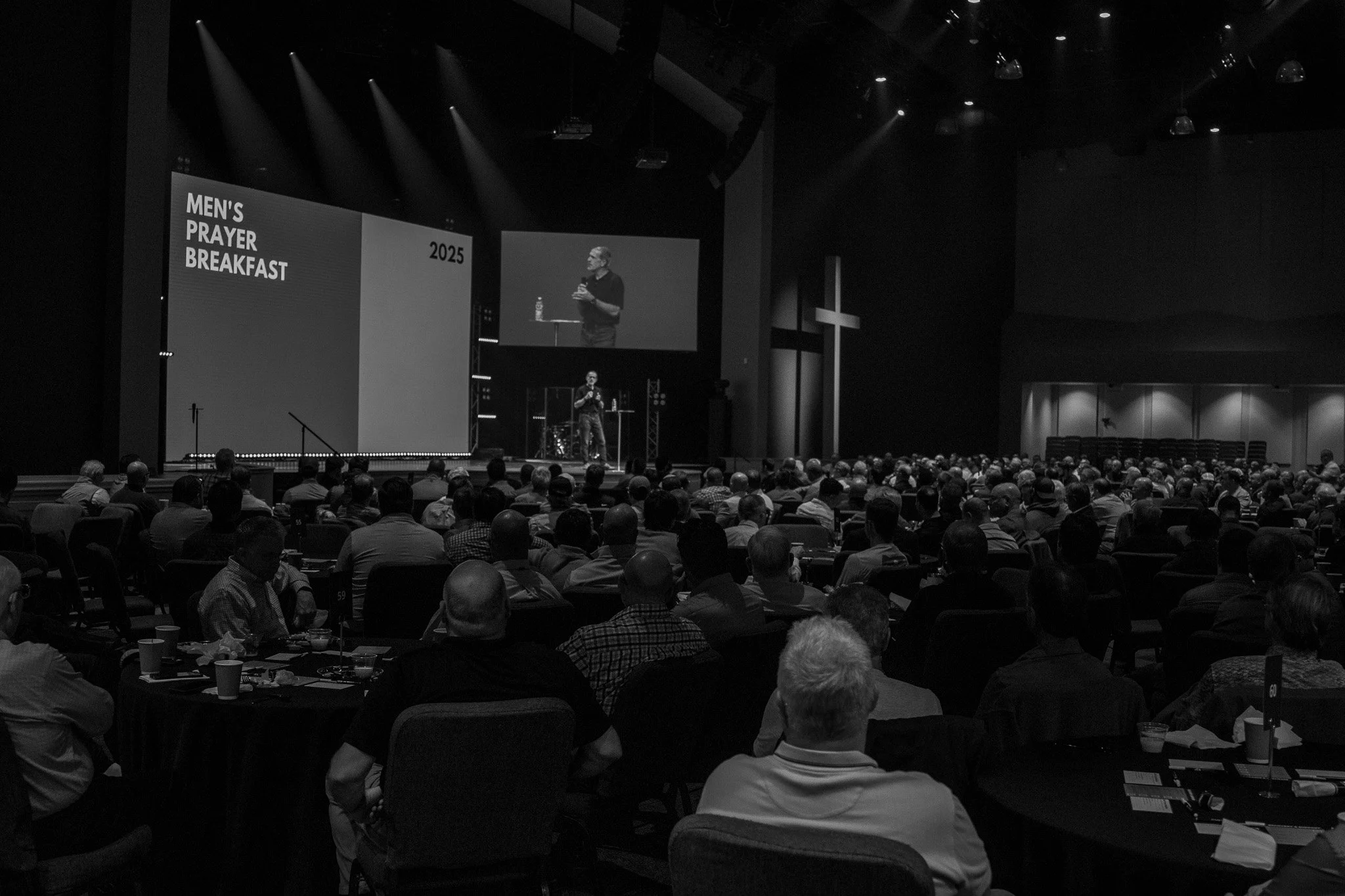 A large indoor church gathering with many attendees seated at tables, a speaker on stage with a microphone, and two large screens displaying the speaker with cross symbols on the sides.