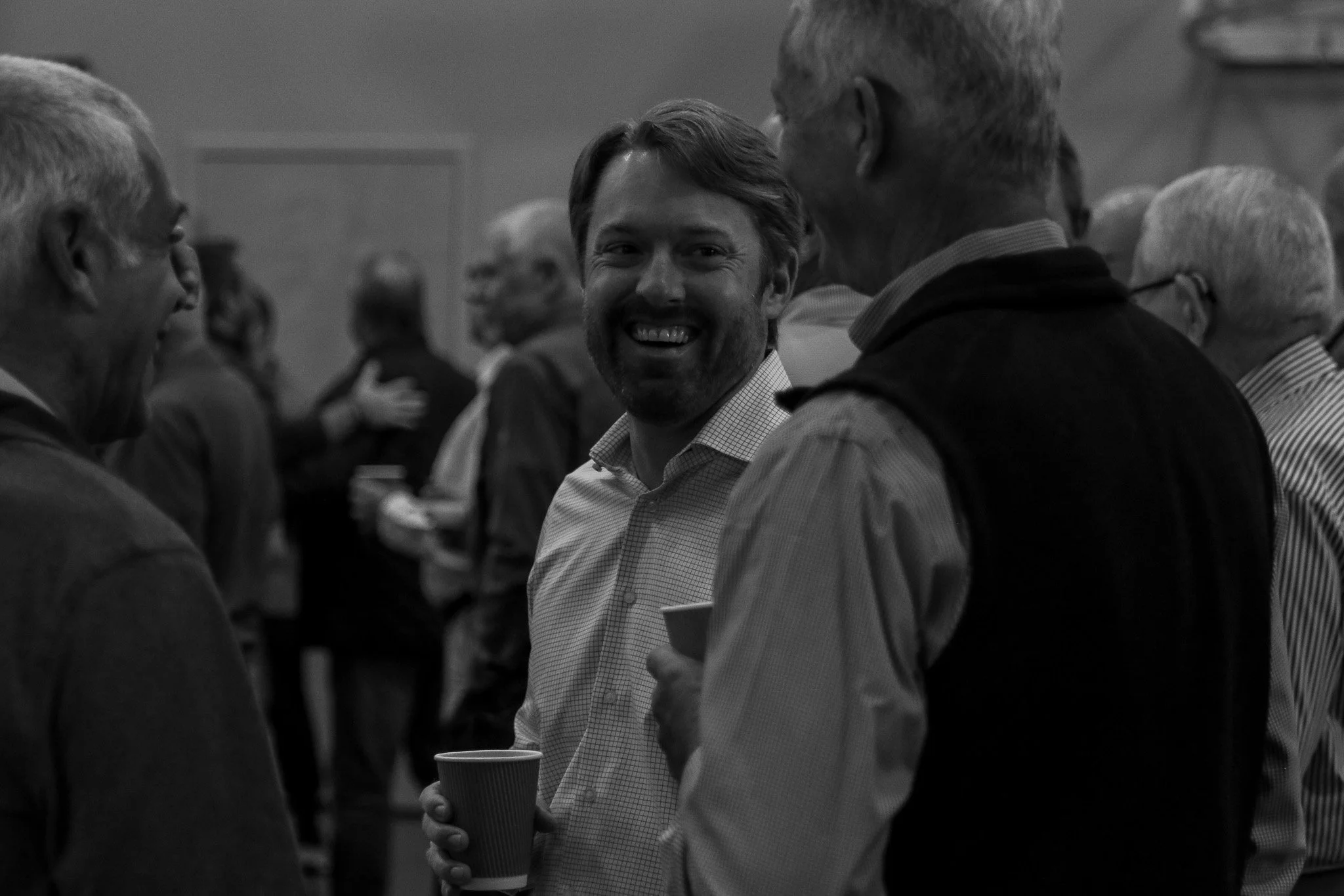 Group of men at a social gathering, one man smiling and holding a paper cup, engaged in conversation.