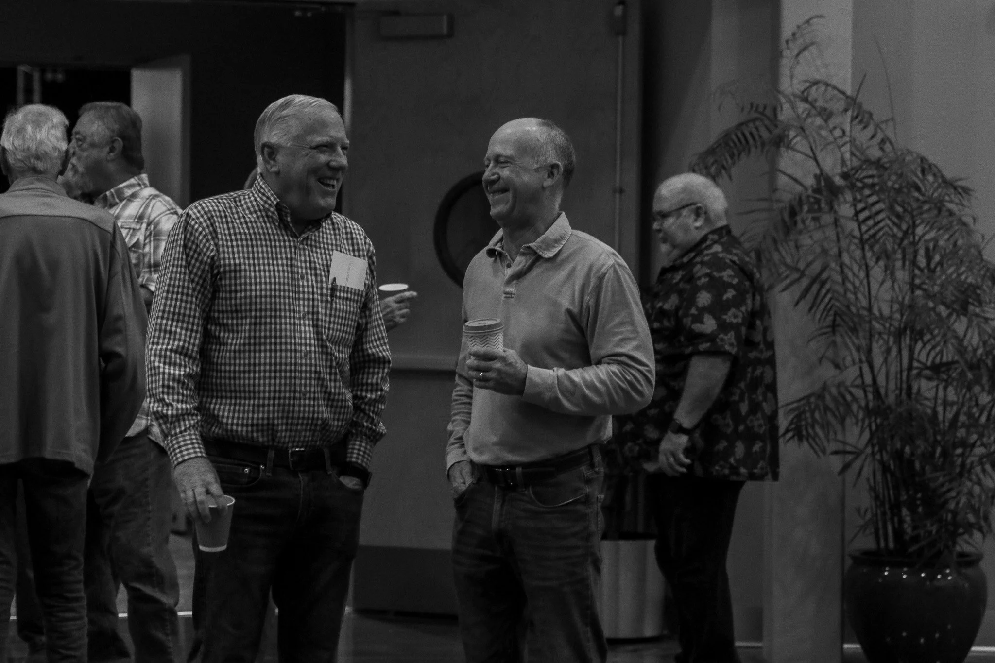 Two men smiling and talking at a social gathering, holding cups, with other people in the background.