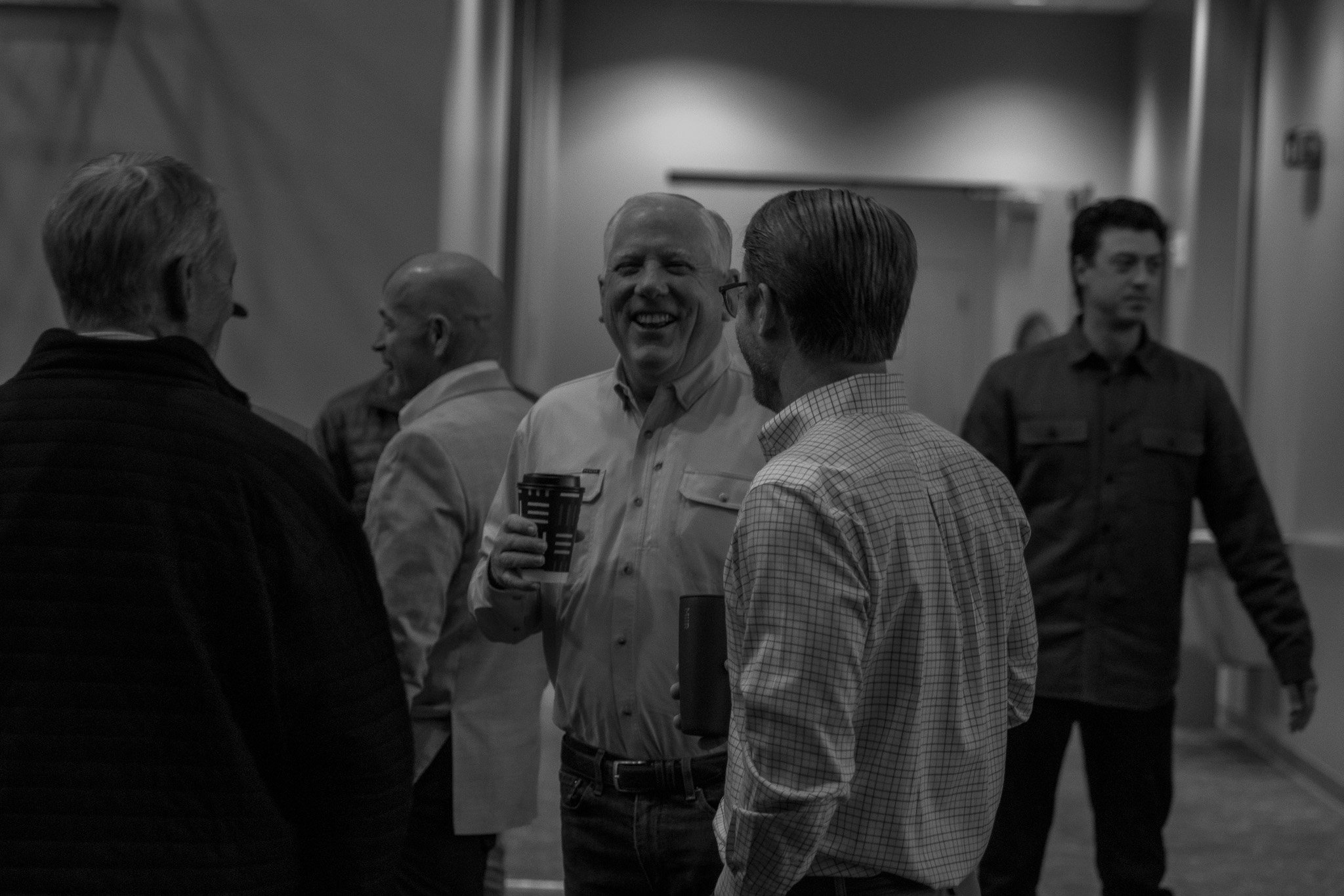 Group of men conversing, smiling, and holding coffee cups in an indoor setting.