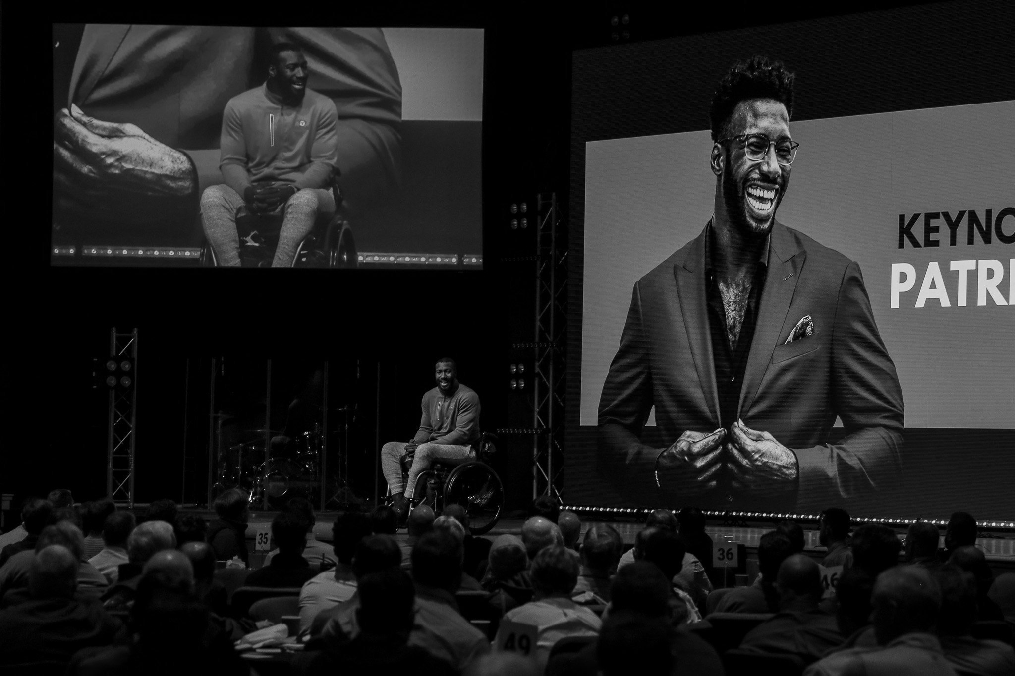 An indoor conference with a large audience and a stage featuring a speaker and large screens displaying his image. The speaker is a man in a wheelchair with glasses and a suit, smiling confidently.