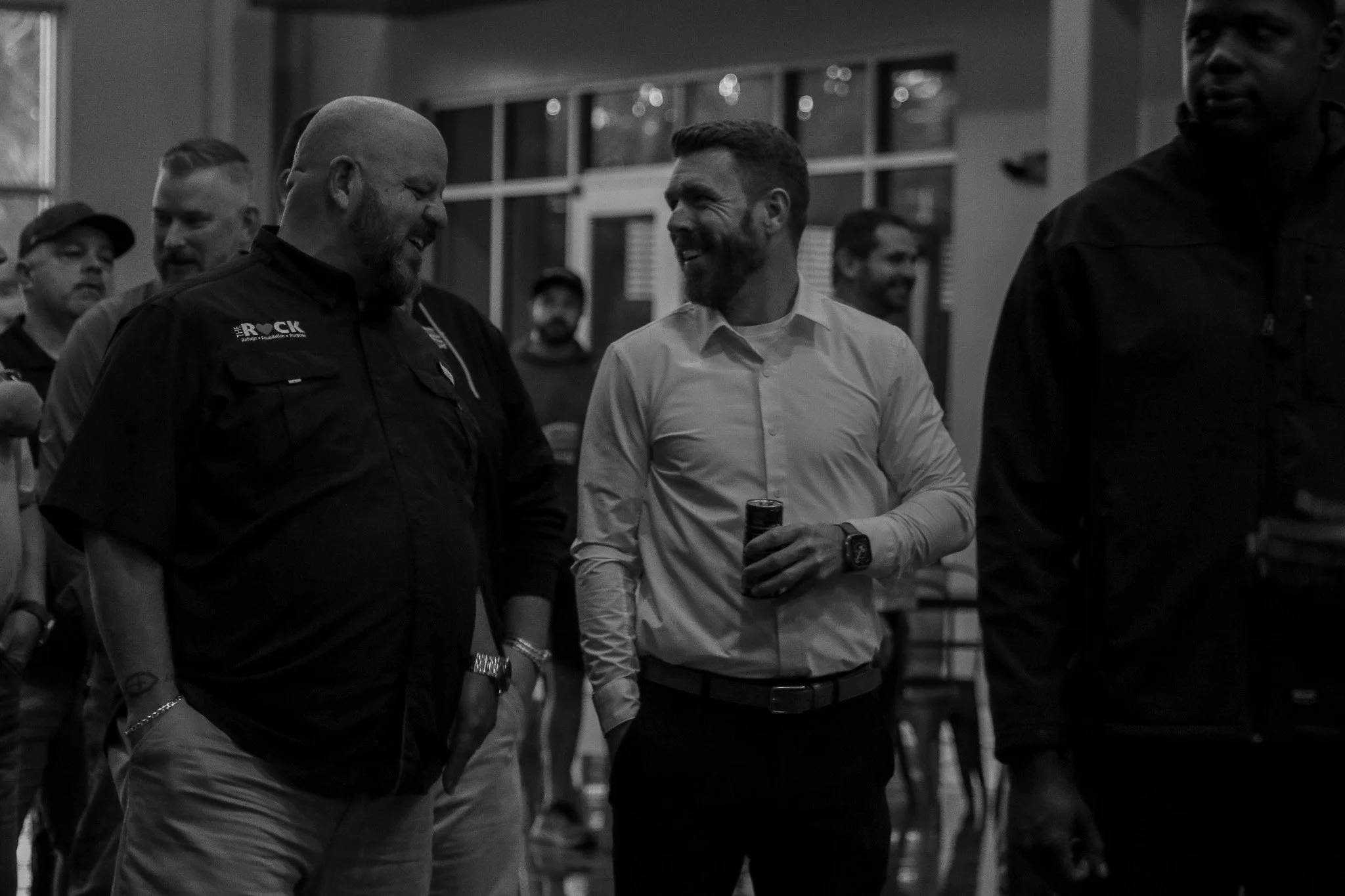 A group of men conversing and smiling indoors, with large windows in the background.