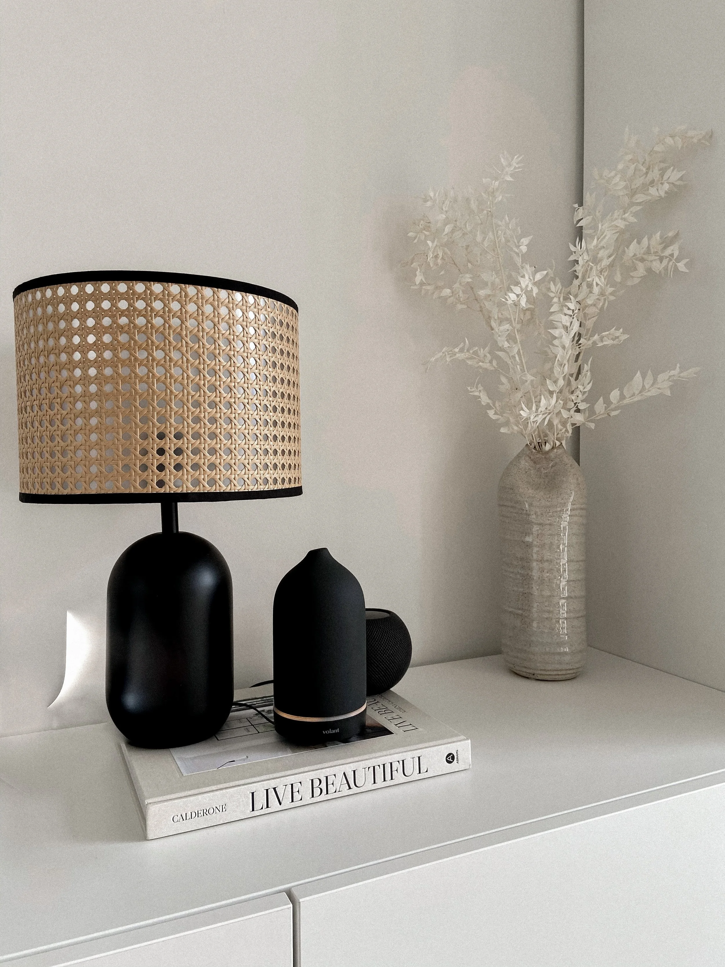 A white table with decorative objects including a black table lamp with a woven rattan shade, two black speakers, a black cylindrical object, and two stacked books titled 'LIVE BEAUTIFUL' and 'LIVRE BEAUTIFUL'. A white ceramic vase with dried white leaves is in the background.
