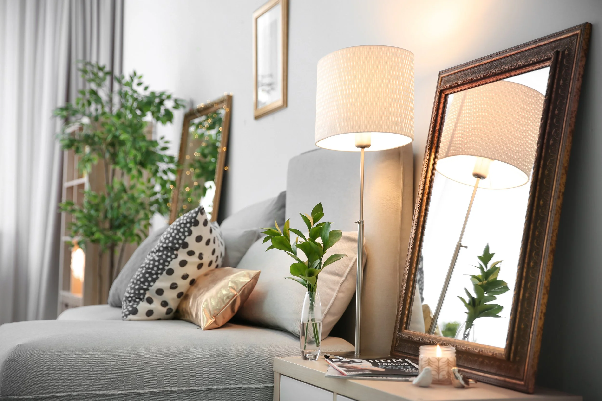 Decorative, cozy bedroom setup with a bed featuring polka dot pillows, a tall lamp, framed mirrors, a potted plant, and candles on a nightstand.