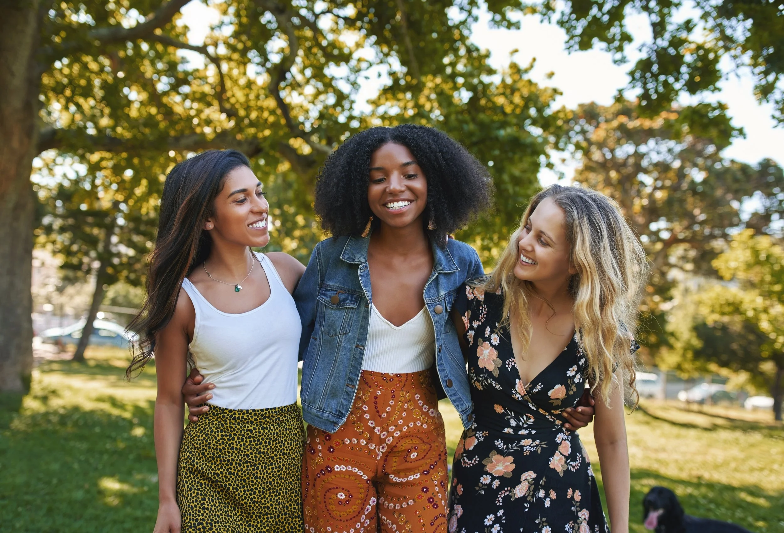 Three women are walking outdoors under trees, smiling and hugging each other.