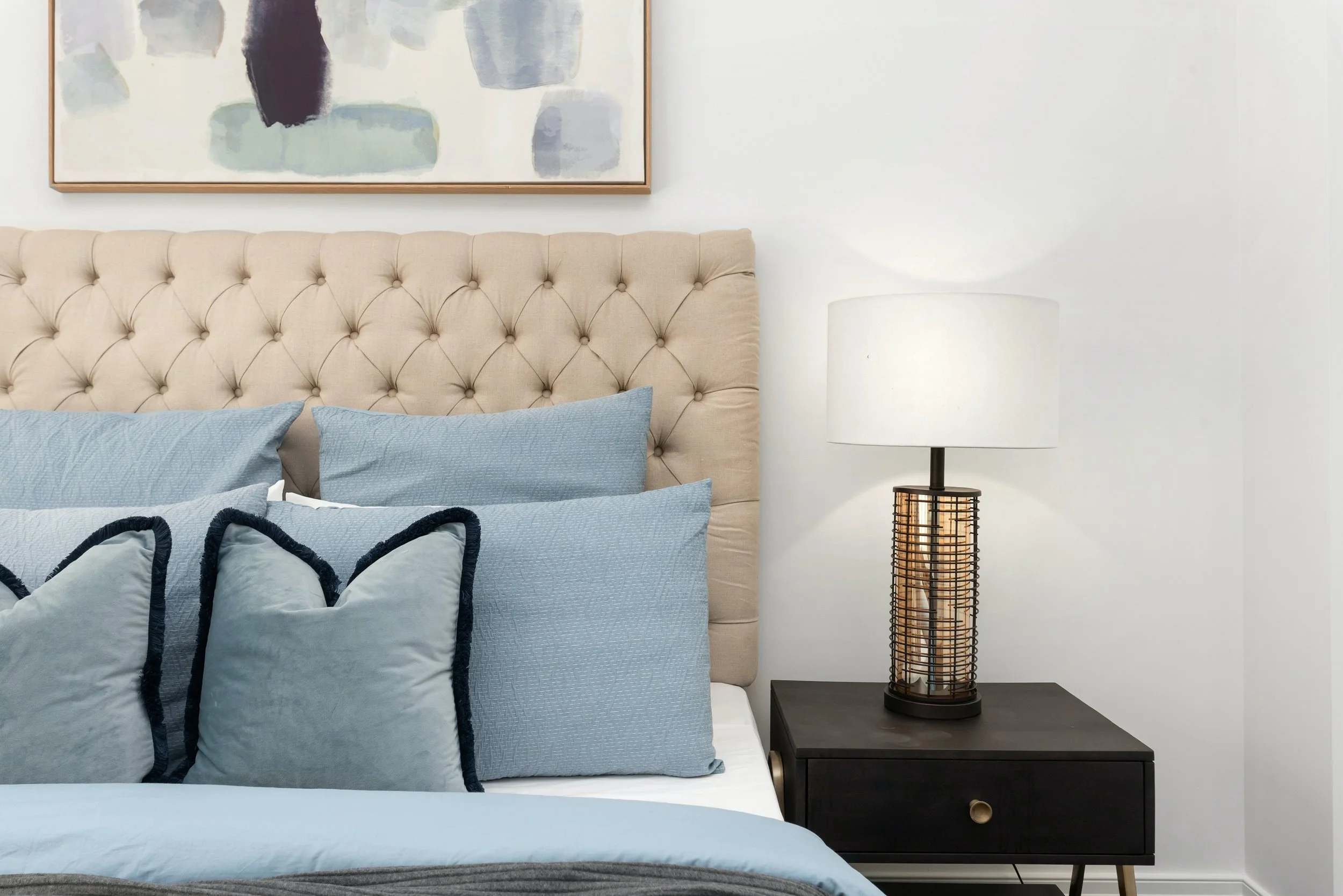 A bedroom with a beige tufted headboard, blue pillows, and a black nightstand with a modern table lamp, and a framed abstract art piece hanging above the bed.