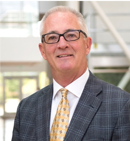 Professional man in glasses, gray blazer, white shirt, and yellow patterned tie, smiling indoors.