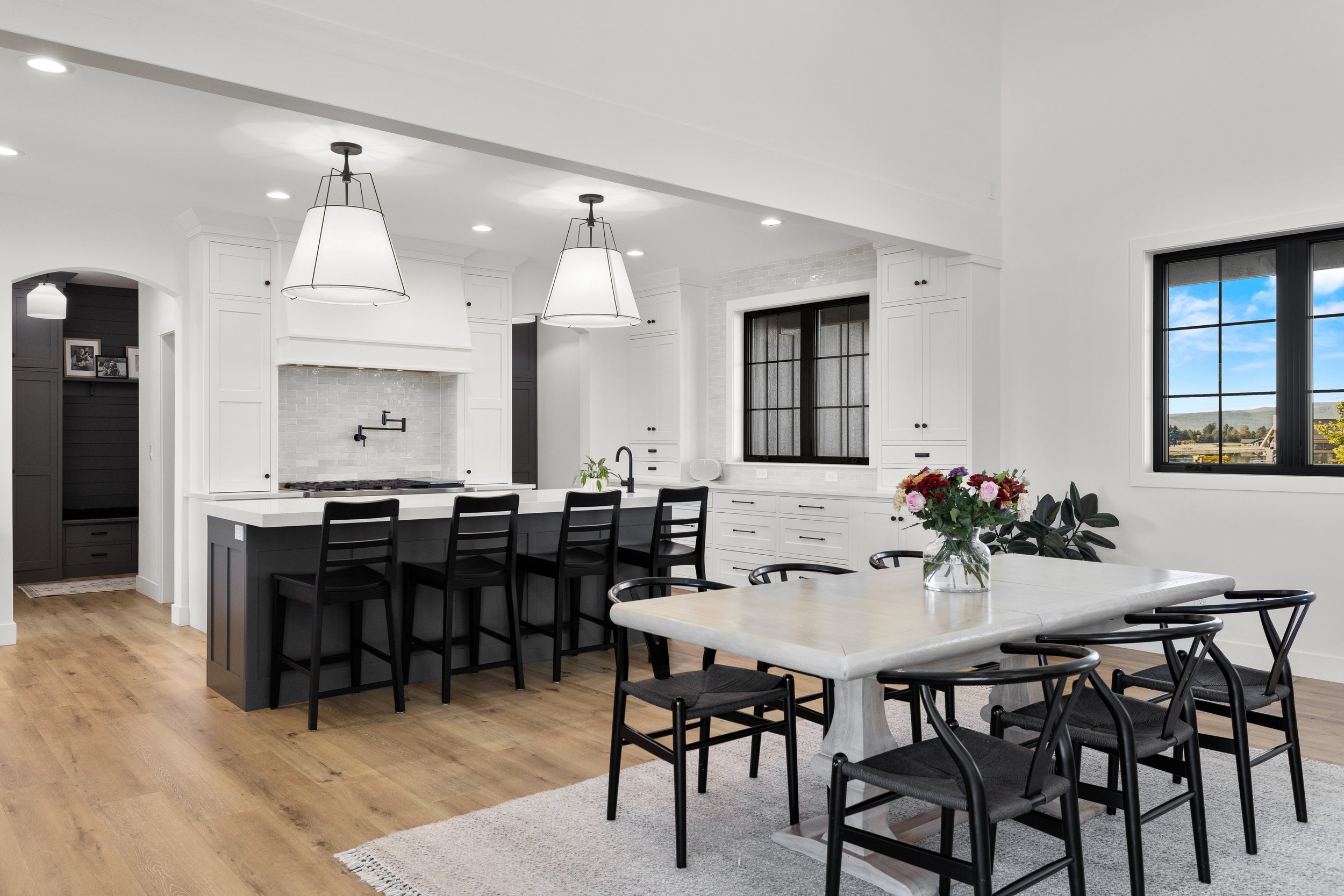 Modern kitchen with black and white cabinets, a large white island, black chairs, two large pendant lights, and a window showing blue sky and landscape.