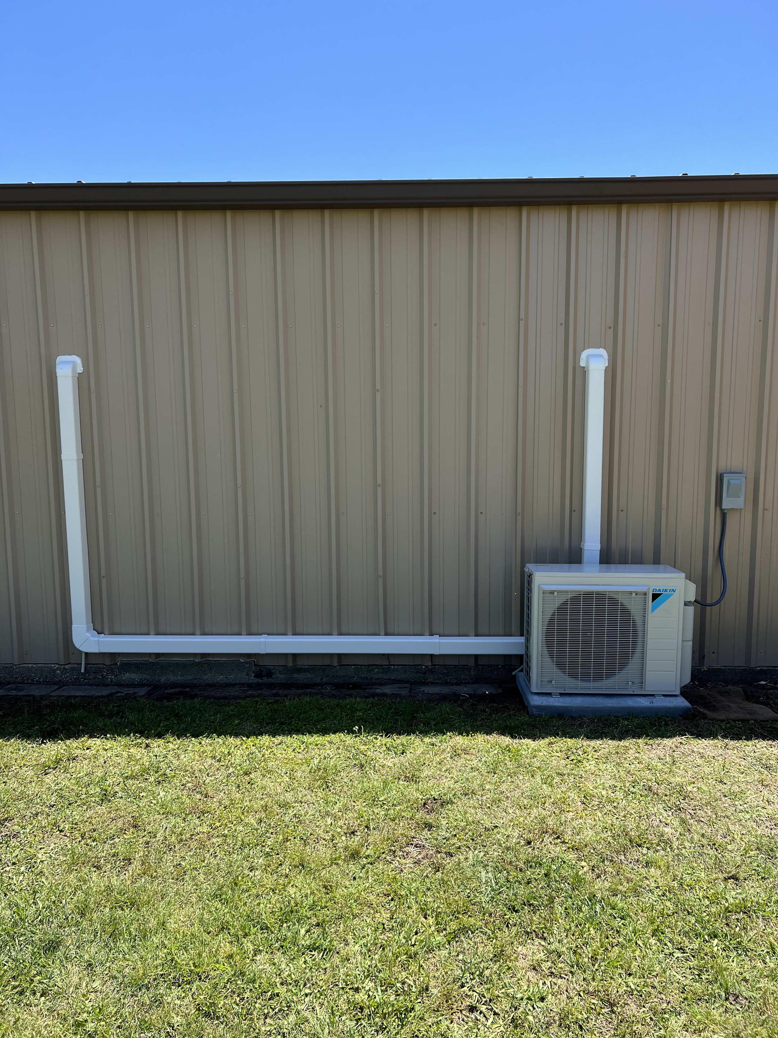The image shows the exterior of a building with beige vertical metal siding, two white pipes running vertically along the wall, an air conditioning unit at the bottom right corner, and a blue sky with no clouds above.
