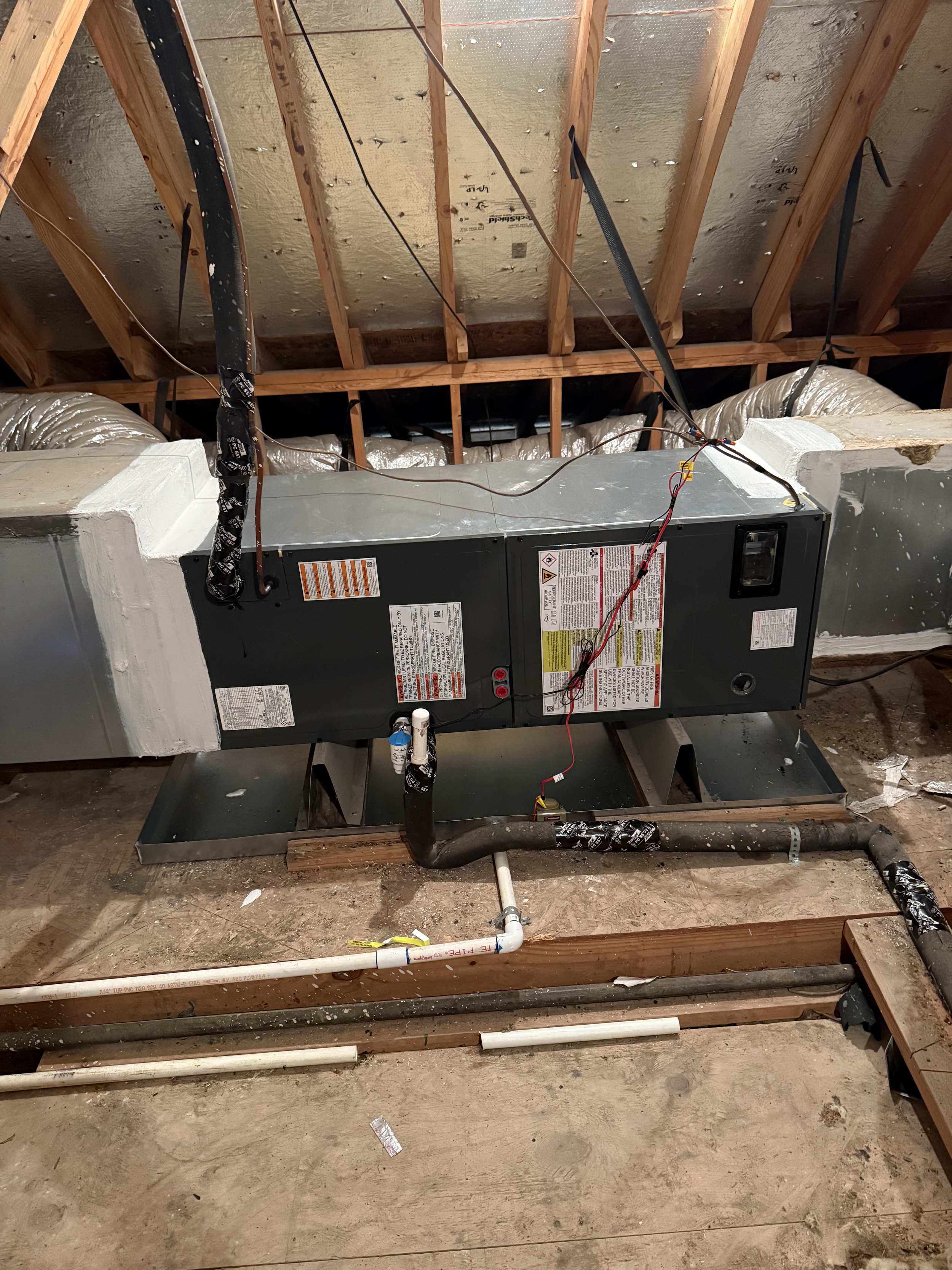 A construction site showing an unfinished ceiling with exposed wooden beams and metal roofing. An HVAC unit is installed below the ceiling, with various wires and pipes connected to it. The floor is dusty with some construction debris.