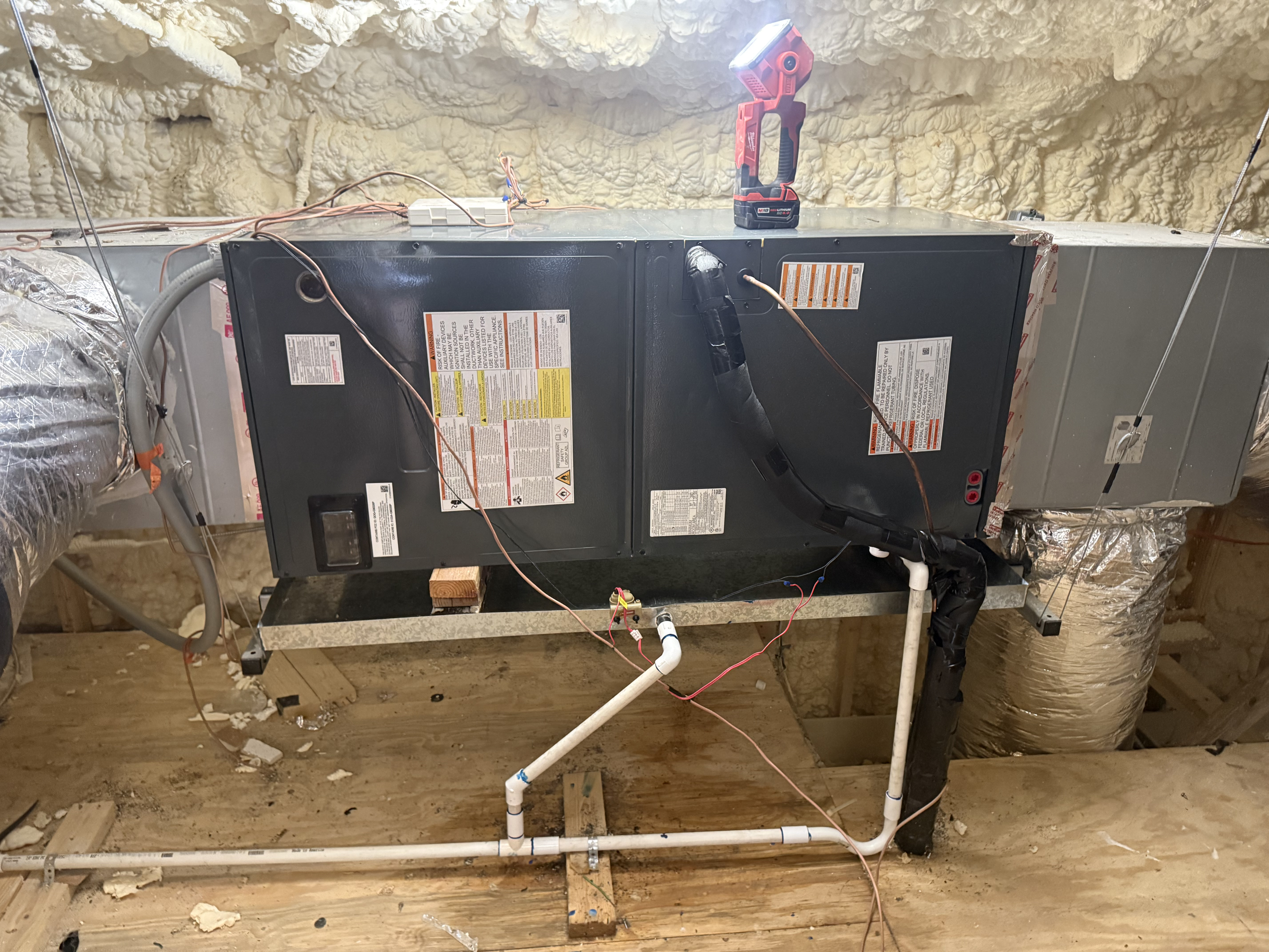 The image shows an HVAC system installed in an attic, with pipes and wires connected to it, surrounded by insulation foam and wood framing.