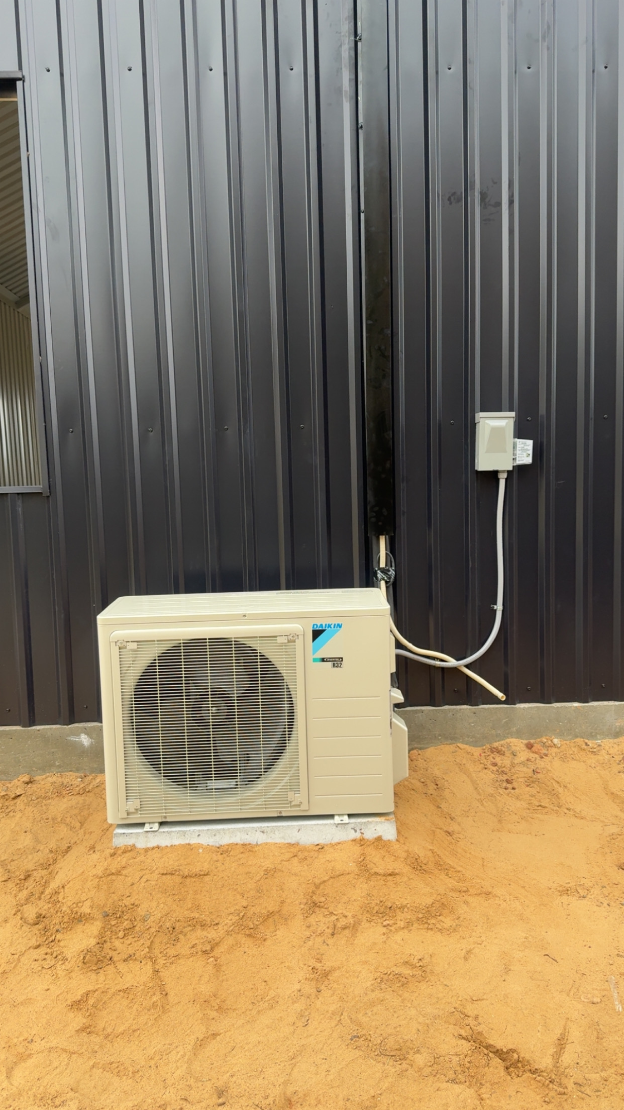 An outdoor air conditioning unit installed on sandy ground in front of a black metal wall with vertical panels.