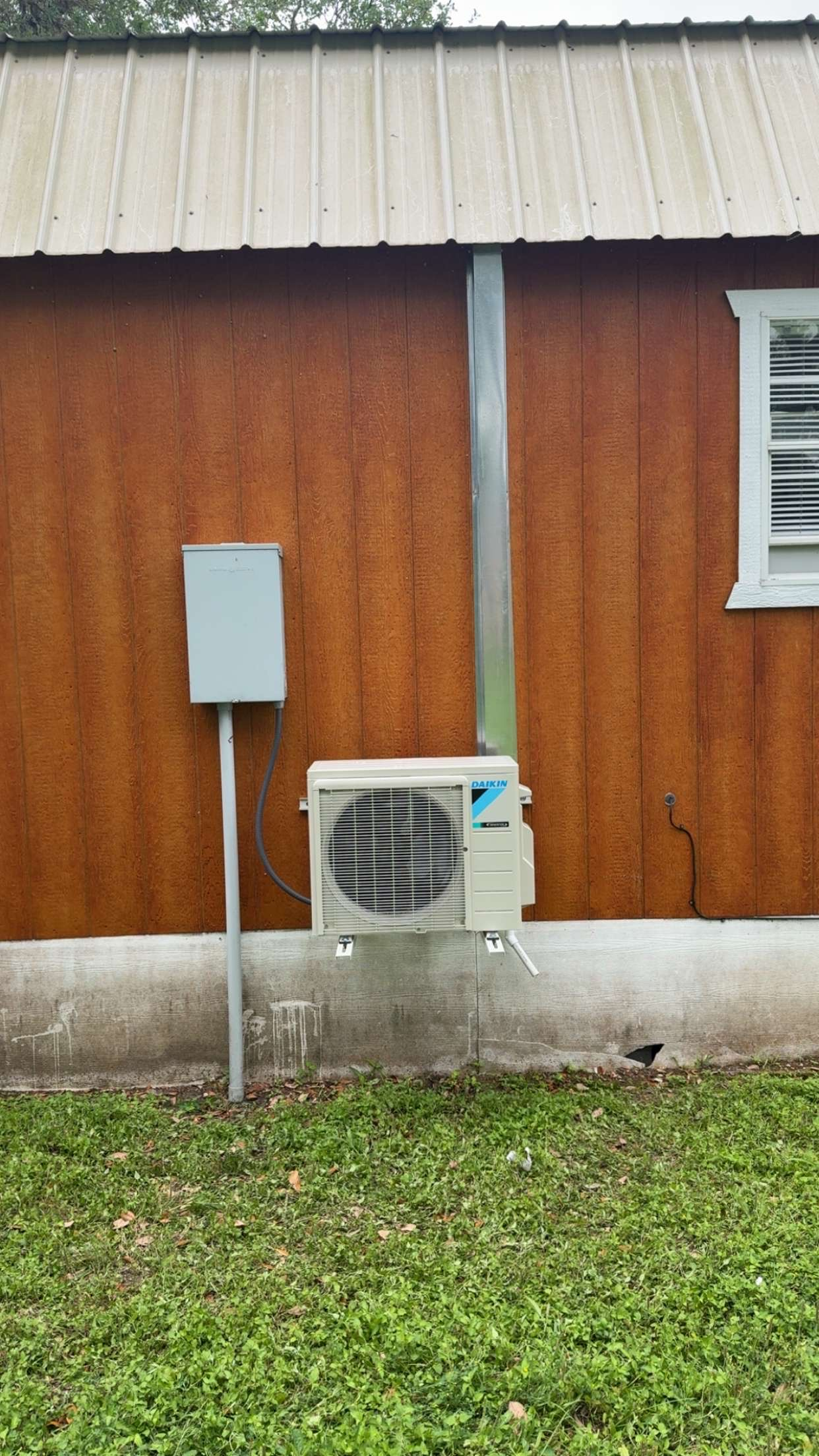 Exterior wall of a house with brown wood siding, a window with blinds, and an air conditioning unit mounted near the ground. A metal pipe runs vertically along the wall, connected to an electrical box.