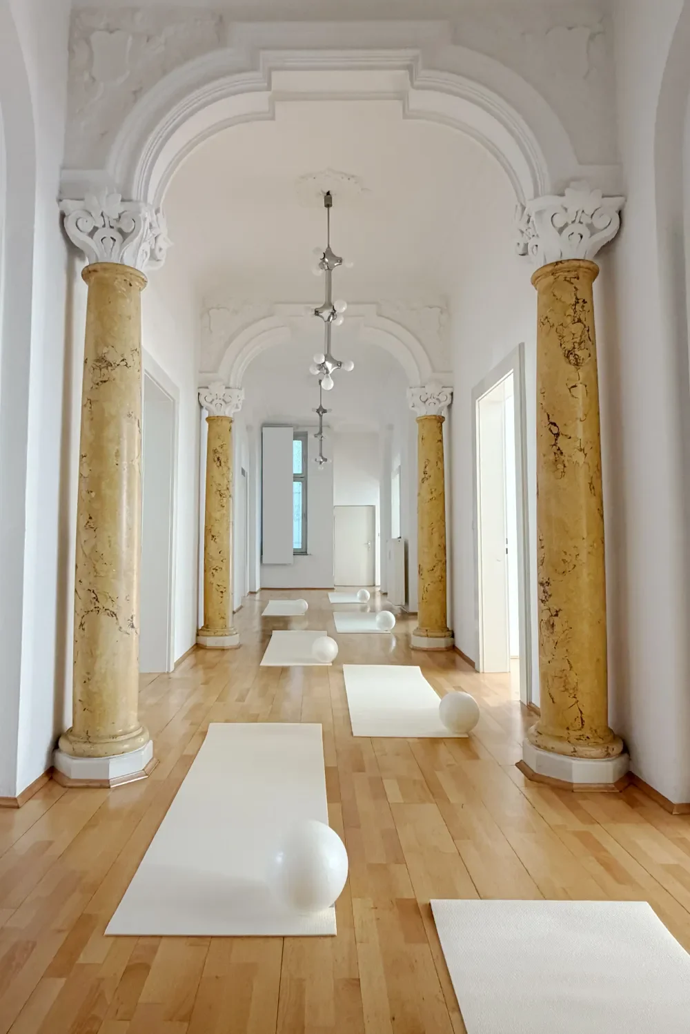 A spacious, elegant corridor with classical architectural design, featuring tall marble columns with ornate capitals, wooden flooring, and white walls. There are white yoga mats with large white exercise balls on top, arranged along the corridor floor. The ceiling has intricate moldings and a modern chandelier.