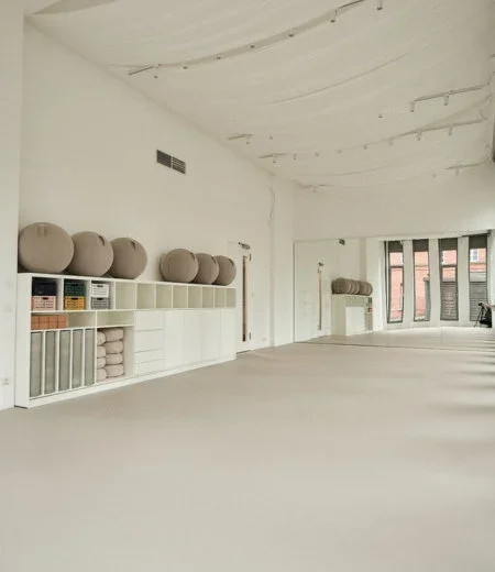 Empty yoga studio with white walls, large windows, and round cushions on white shelves.