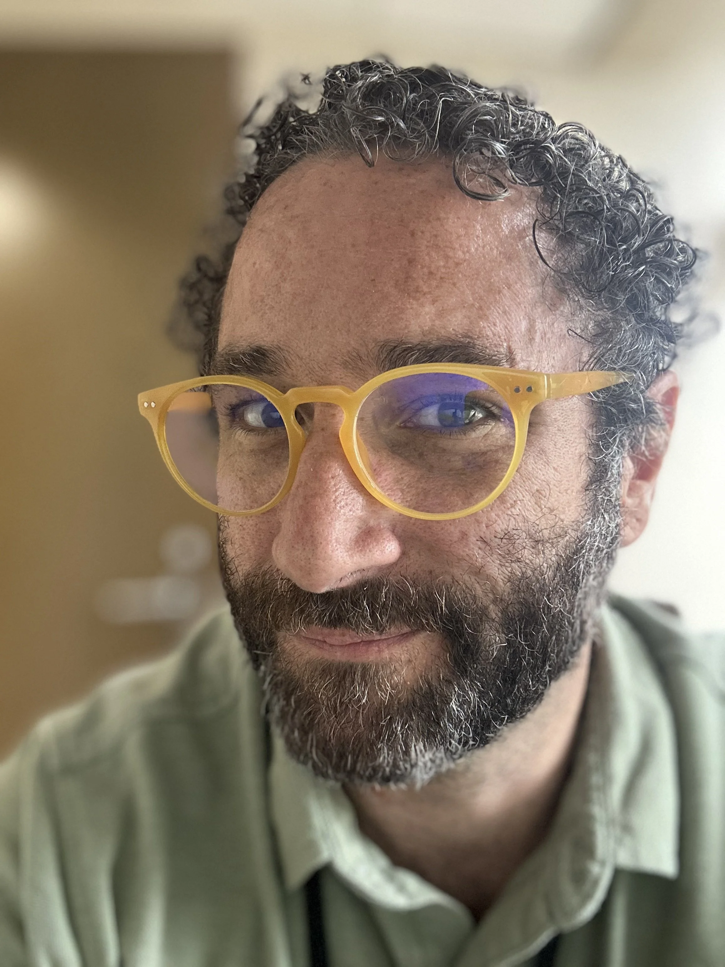 A close-up of a man's face partially visible, wearing yellow glasses with purple lenses and having curly dark hair and a beard, with a blurred background.
