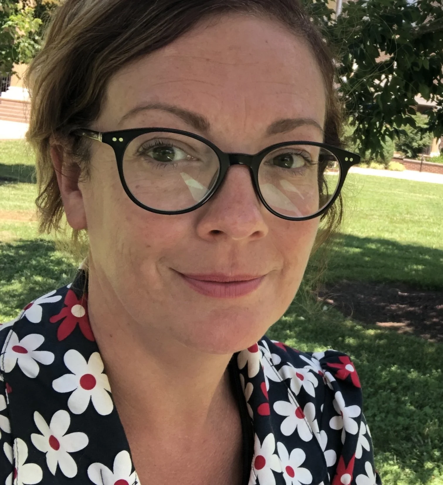 Close-up of a woman wearing black glasses with a floral patterned dress, outdoors with green lawn and trees in the background.