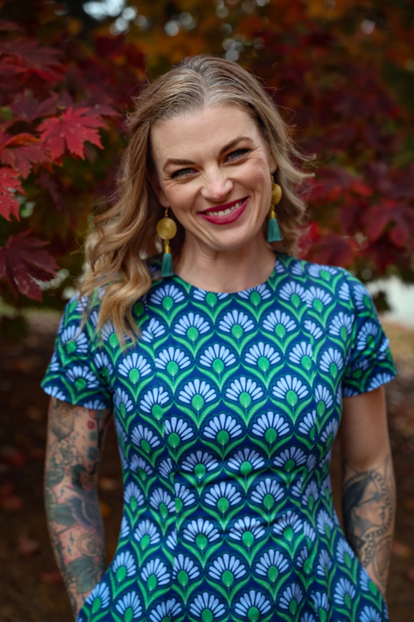 A woman with wavy, shoulder-length blonde hair smiling outdoors. She is wearing a colorful dress with a blue, green, and white floral pattern, large gold earrings with teal tassels, and has tattoos on both arms. The background features red and orange autumn leaves.